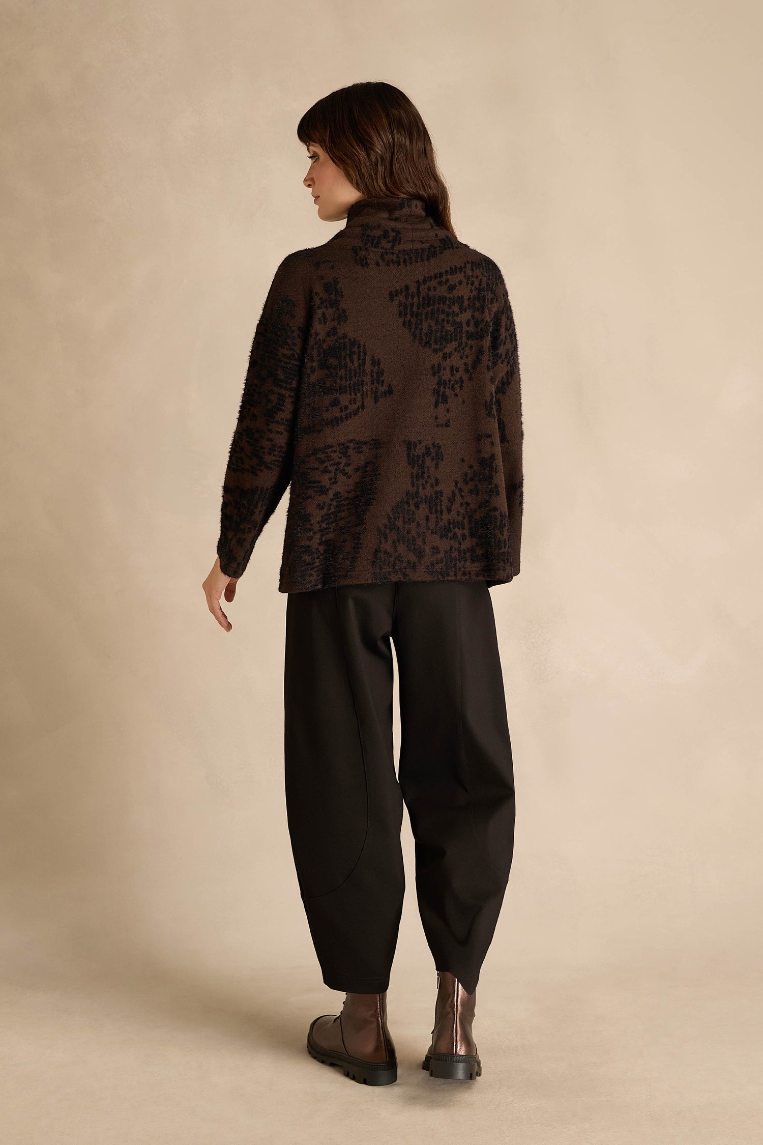 A person stands facing away, wearing the Cocoa Speckled Jersey Boxy Top with a brown abstract pattern and boxy fit, paired with black loose pants and dark ankle boots, against a neutral beige background.