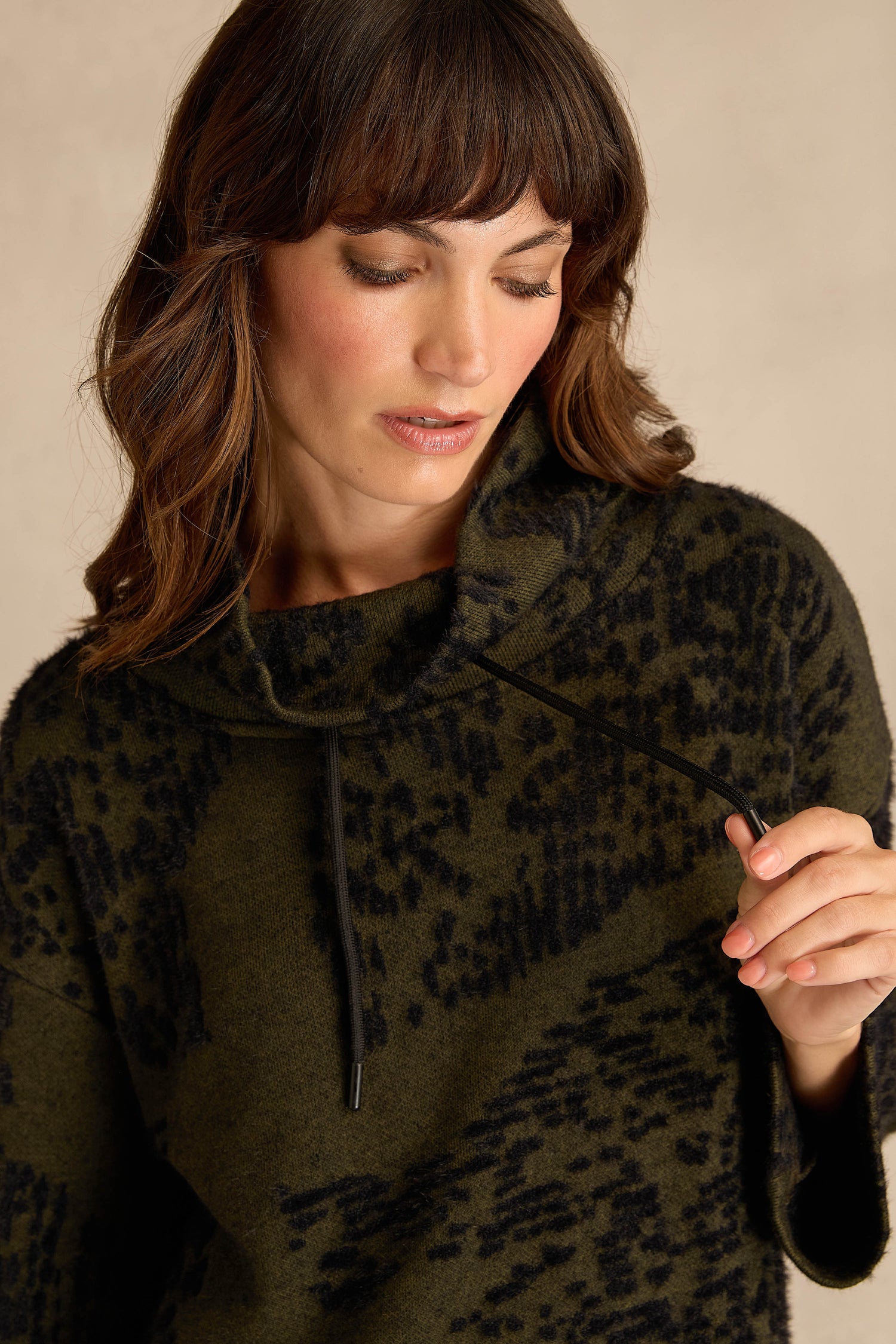 A woman with shoulder-length brown hair wears the Thyme Speckled Jersey Boxy Top, looking down as she holds the top’s drawstring in her right hand.