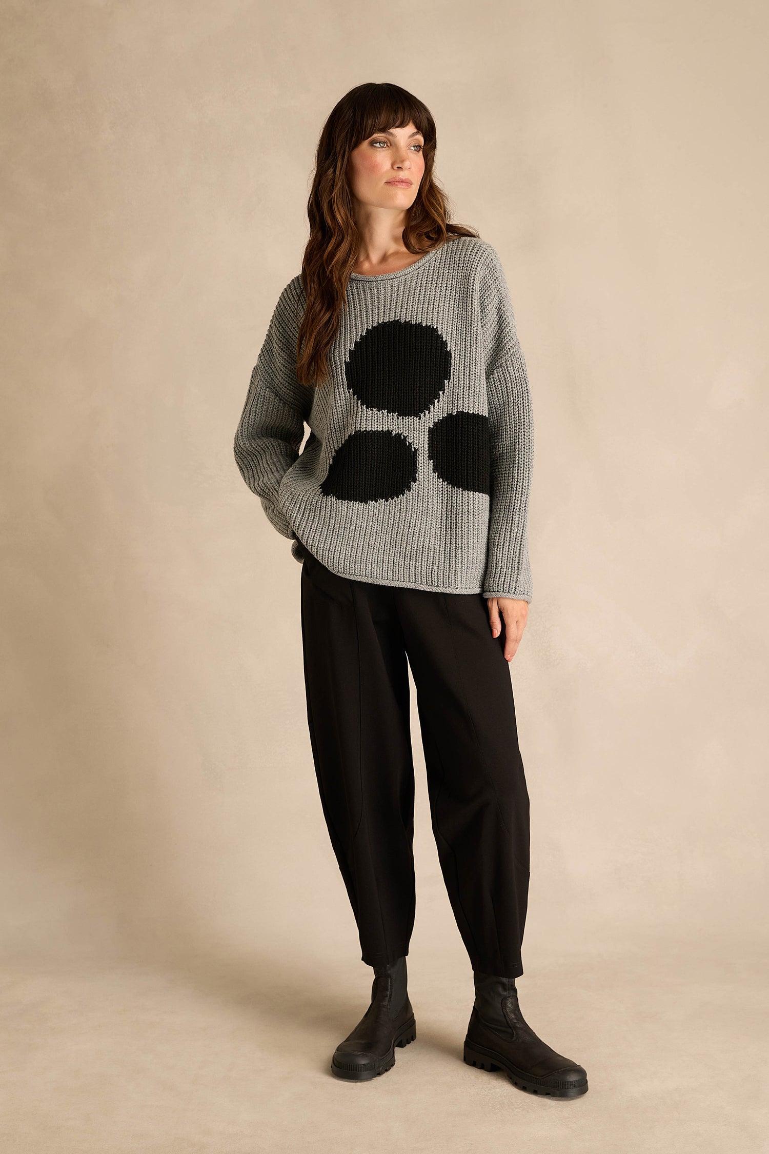 A woman stands against a neutral background, dressed in the Grey Spot Scoop Neck Knit—an alpaca and wool sweater featuring an oversized spot motif with large black circles—paired with black pants and black boots.