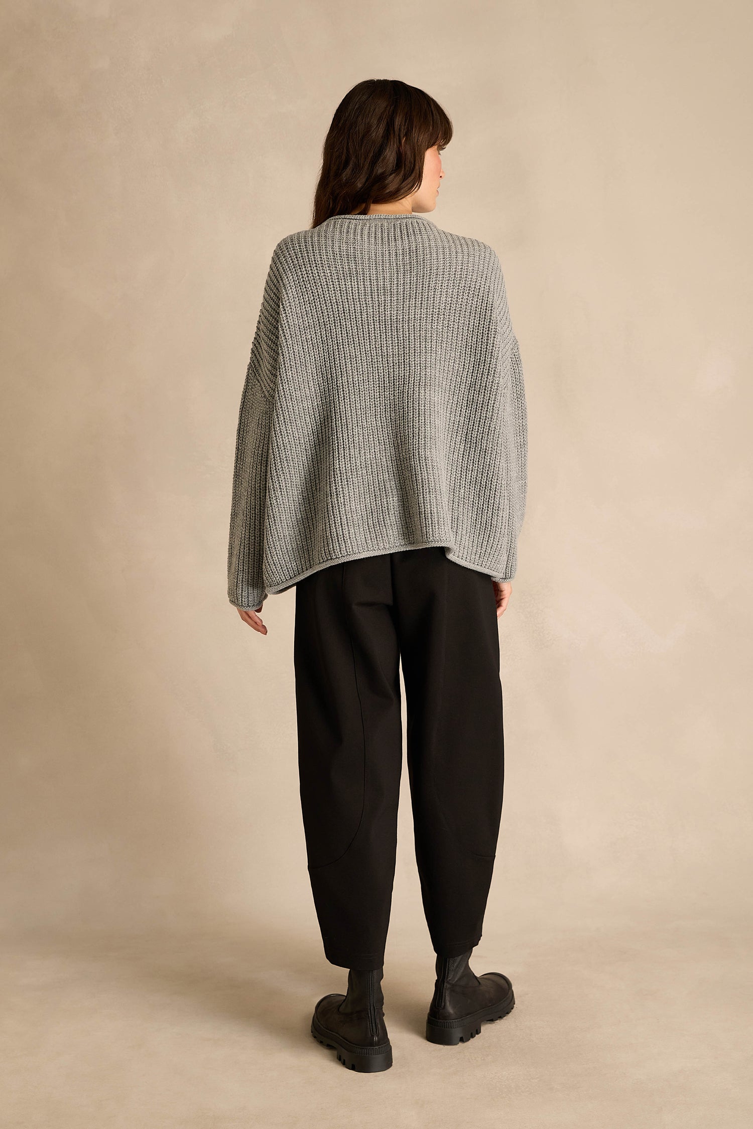 A person stands with their back to the camera, wearing the Grey Spot Scoop Neck Knit—a loose gray sweater crafted from wool and alpaca—paired with black pants and black boots, set against a plain beige background.