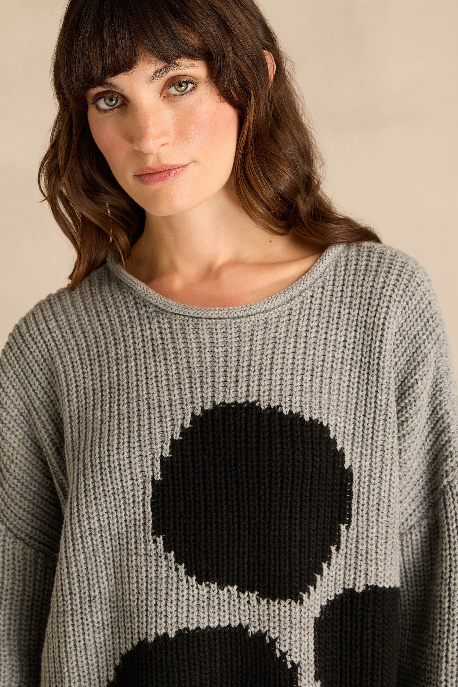 A woman with shoulder-length brown hair and bangs is wearing the Grey Spot Scoop Neck Knit, a grey ribbed-knit sweater crafted from luxurious wool and alpaca, featuring an oversized spot motif and a stylish scoop neckline.