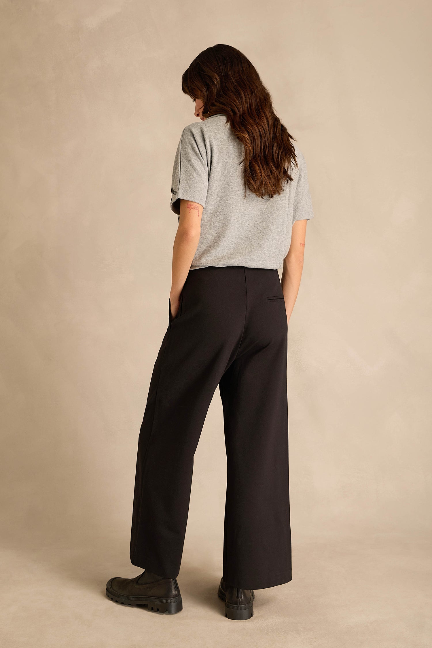 A person stands with their back to the camera, wearing a gray short-sleeve shirt, black shoes, and the Wide Leg Ponte Trousers—black wide leg trousers crafted from stretch ponte jersey—set against a neutral background.