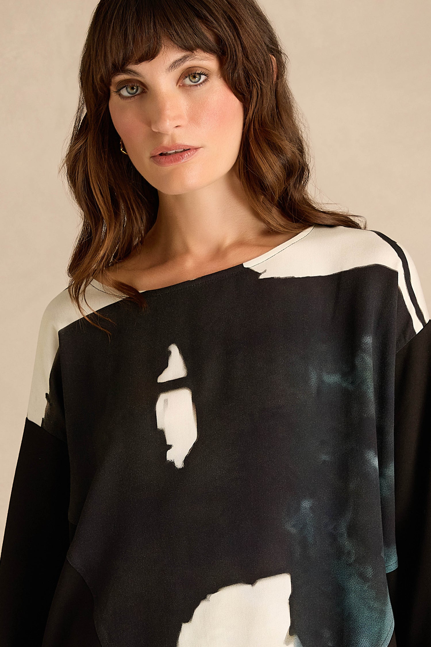 A woman with wavy brown hair is wearing the Abstract Watercolour Scoop Neck Top—a loose, long sleeve, relaxed fit top featuring an abstract pattern—while looking at the camera against a neutral background.