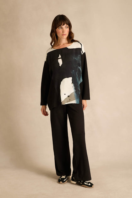 A woman stands against a plain beige background wearing the Abstract Watercolour Scoop Neck Top—a loose-fitting, black and white long-sleeve top with an abstract pattern—paired with black pants and black slip-on shoes.