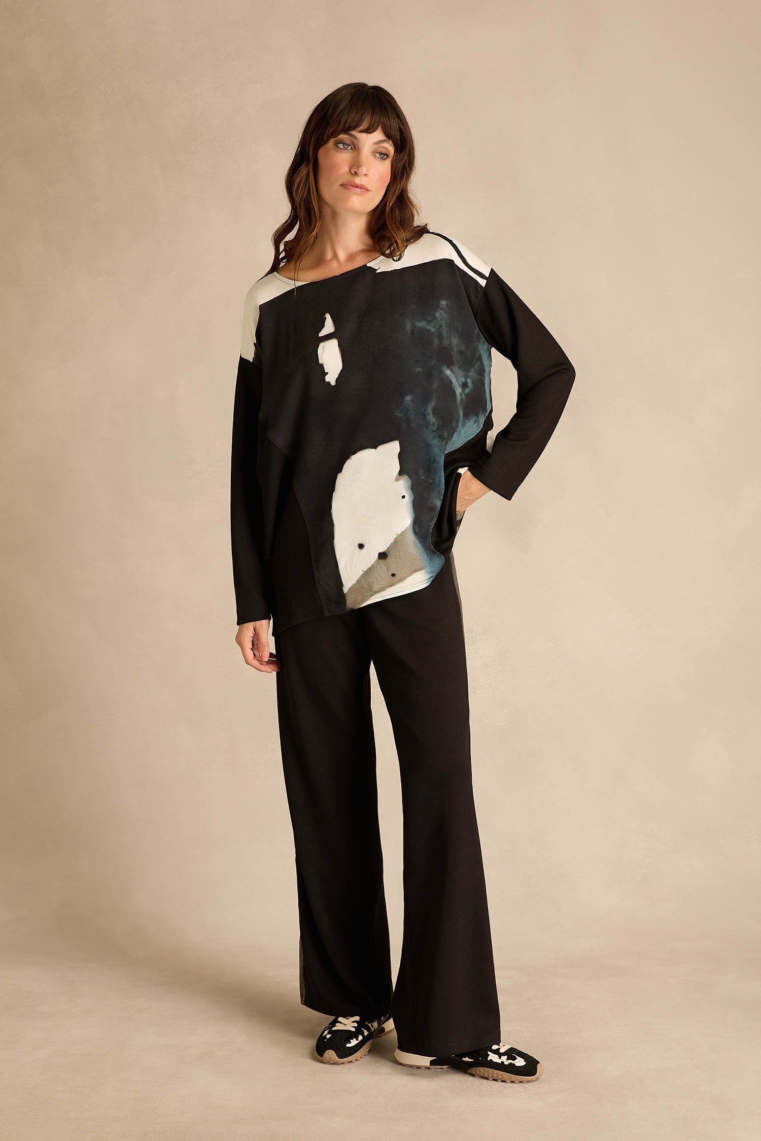 A woman stands facing forward against a neutral studio backdrop, wearing black wide-leg pants, black loafers with white detailing, and the Abstract Watercolour Scoop Neck Top—a long sleeve relaxed fit top.