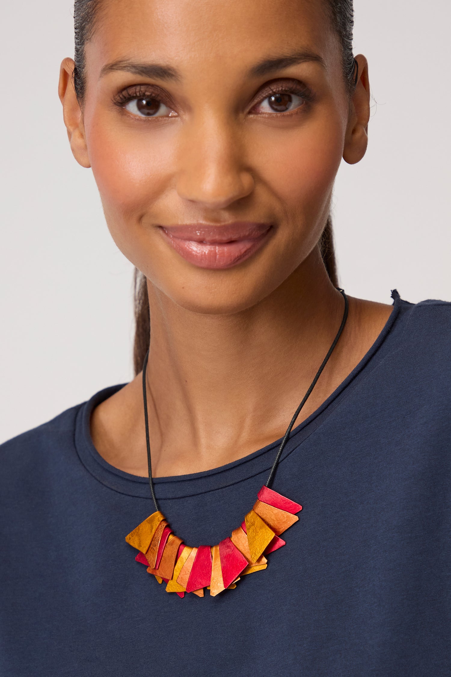 A woman in a navy top smiles at the camera, wearing the Mini Segment Necklace—an Oana Millet design with geometric red and yellow accents—against a neutral background.
