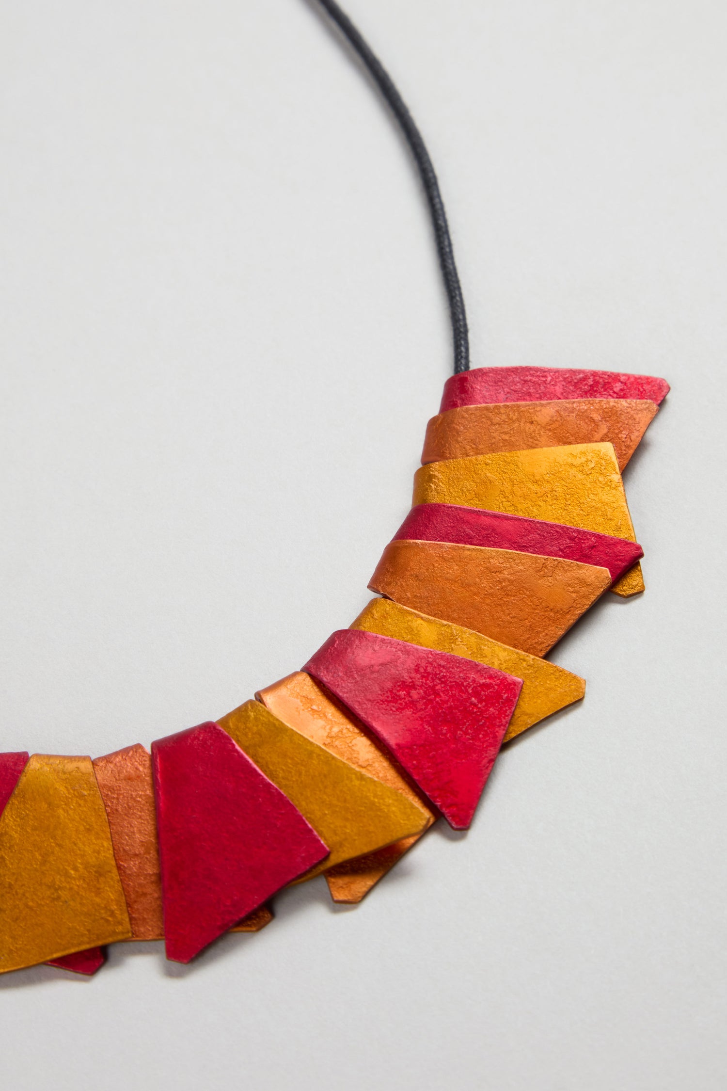 A close-up of the Mini Segment Necklace showcases geometric red and orange shapes on a black cord, making it a striking example of contemporary jewellery displayed against a light background.