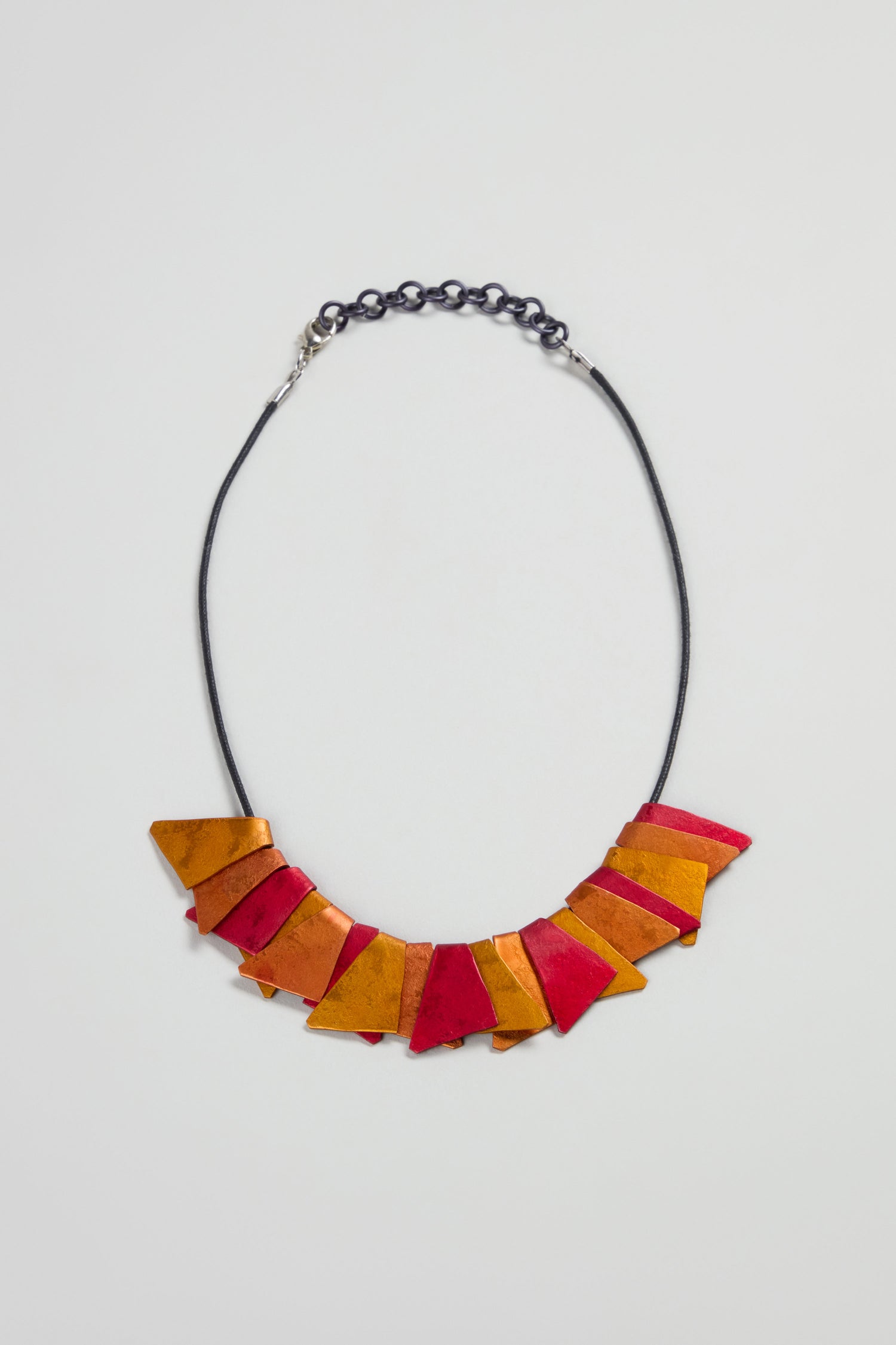 The Mini Segment Necklace features a black cord and chain with alternating orange and red triangles, offering a bold modern look.