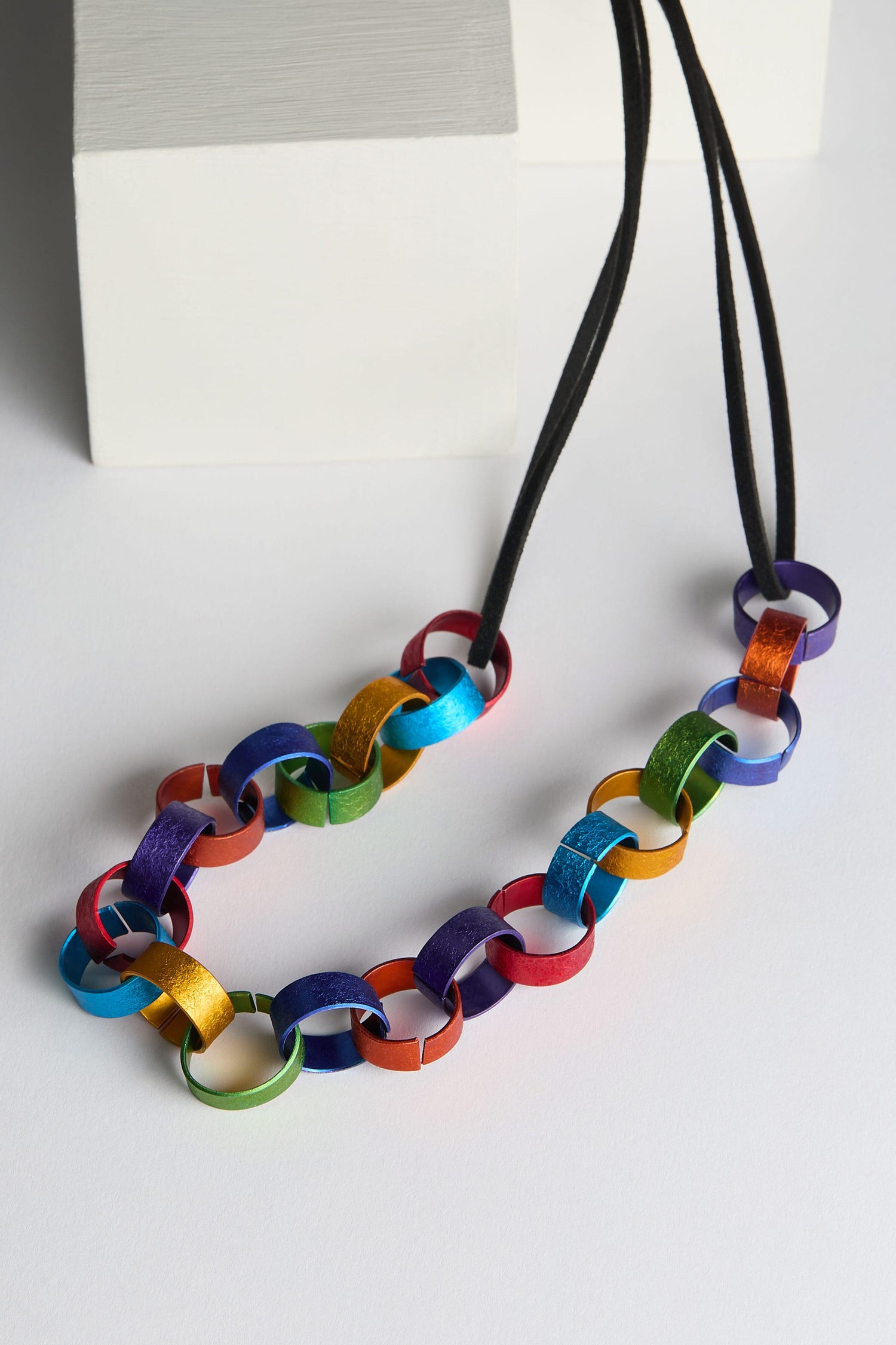 The Rainbow Links Necklace, featuring multicolored metal links and a black adjustable cord, displayed against white geometric shapes, makes for stunning statement jewelry.