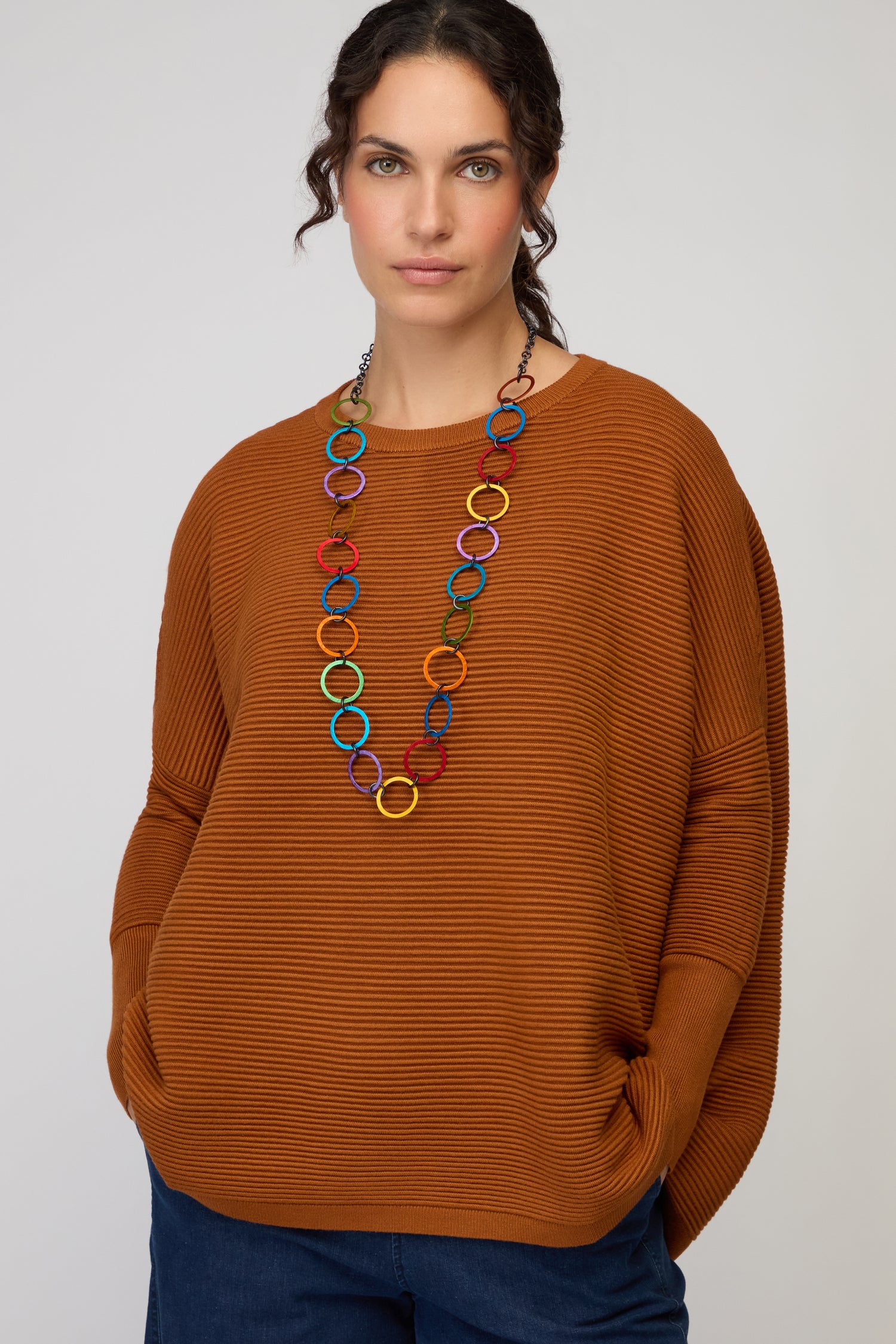 A woman stands with hands in pockets against a plain background, wearing a rust ribbed sweater, dark blue pants, and the Rainbow Rings Necklace—an anodised aluminium piece by Oana Millet.