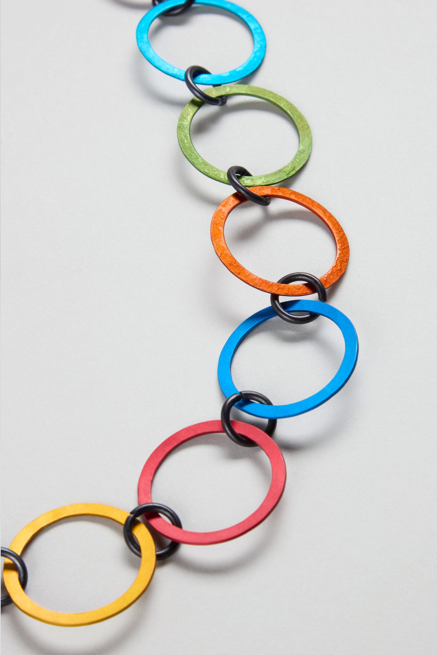 The Rainbow Rings Necklace features interlocking metal rings in blue, green, orange, yellow, and red on a light gray background—vibrant anodised aluminium jewellery by Oana Millet.