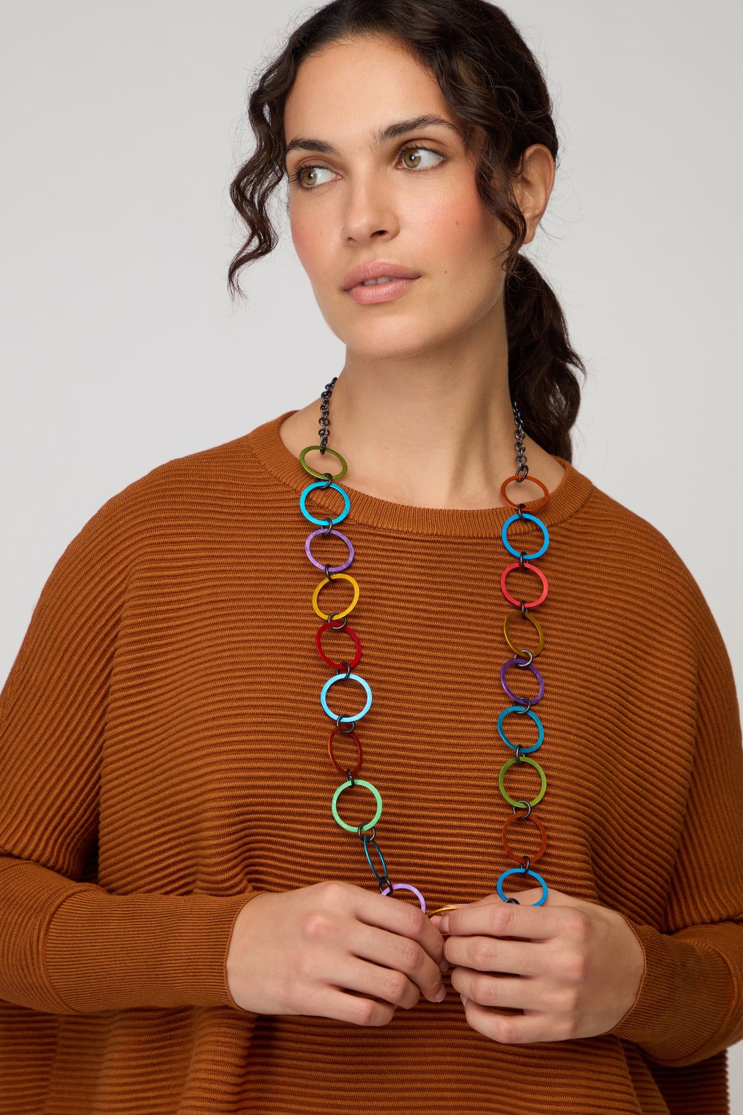 A woman wearing a rust-colored ribbed sweater and the Rainbow Rings Necklace, looking slightly to the side against a neutral background.