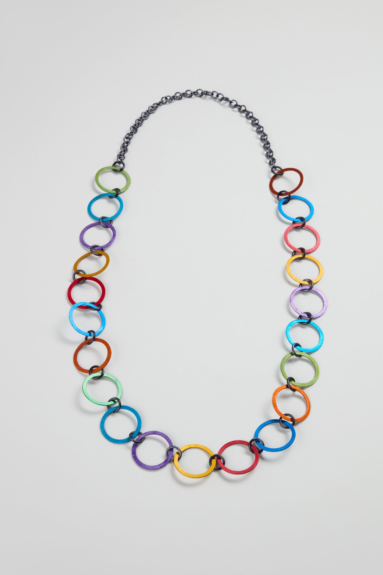 The Rainbow Rings Necklace showcases vibrant, large interlocking anodised aluminium rings with a dark chain section at the top, set against a plain white background.