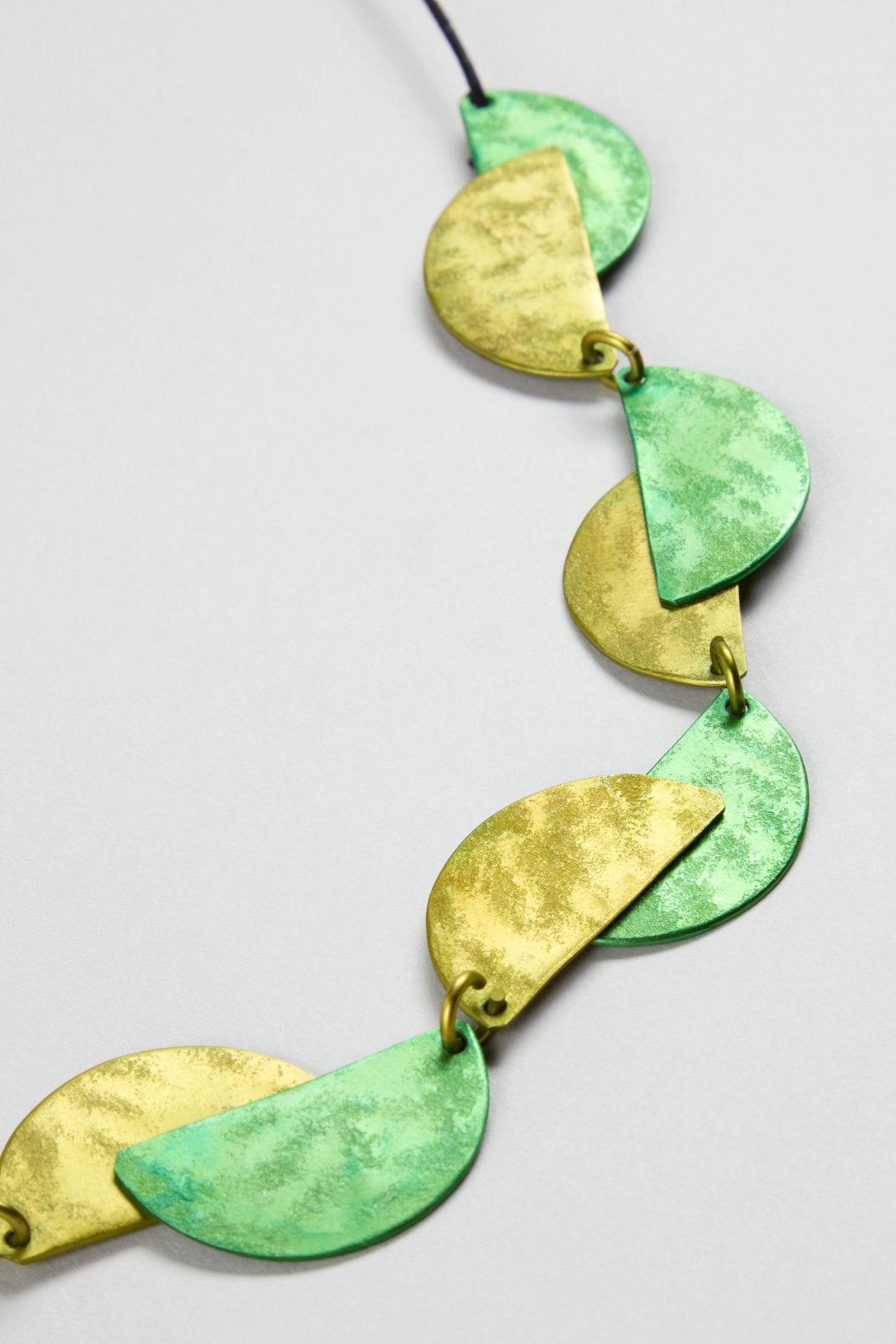 The Half Moon Necklace features alternating green and yellow semi-circular metal pieces linked by small rings in a striking geometric pattern.