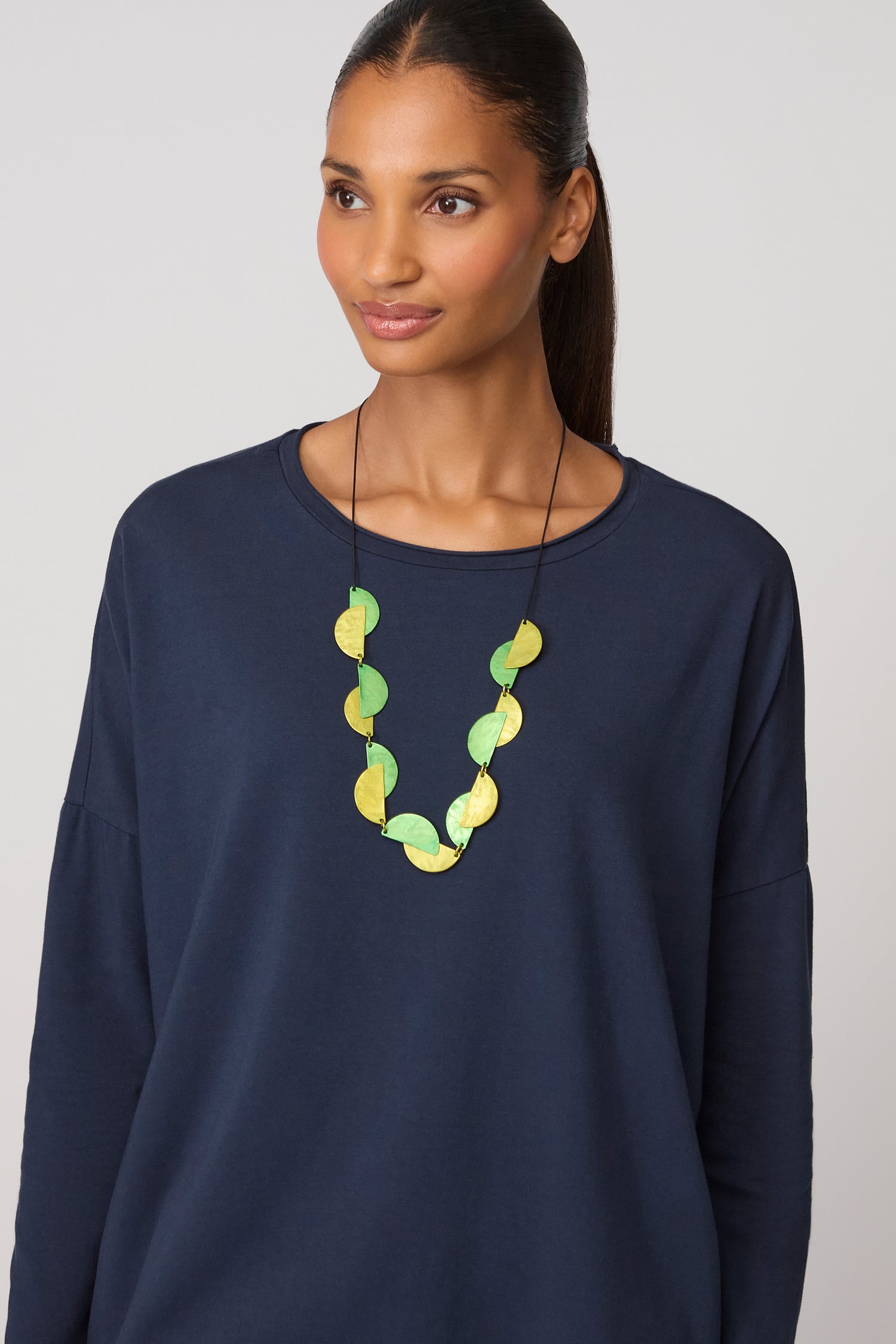 A woman wearing a navy blue long-sleeve top and the Half Moon Necklace, featuring green and yellow geometric circles, stands against a plain light background.