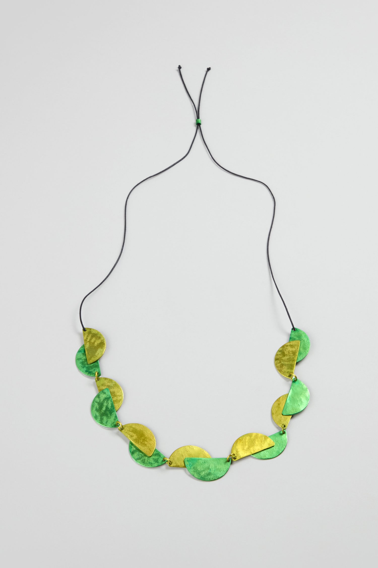 The Half Moon Necklace features geometric green and gold oval pieces on a black adjustable cord, showcased against a light gray background.