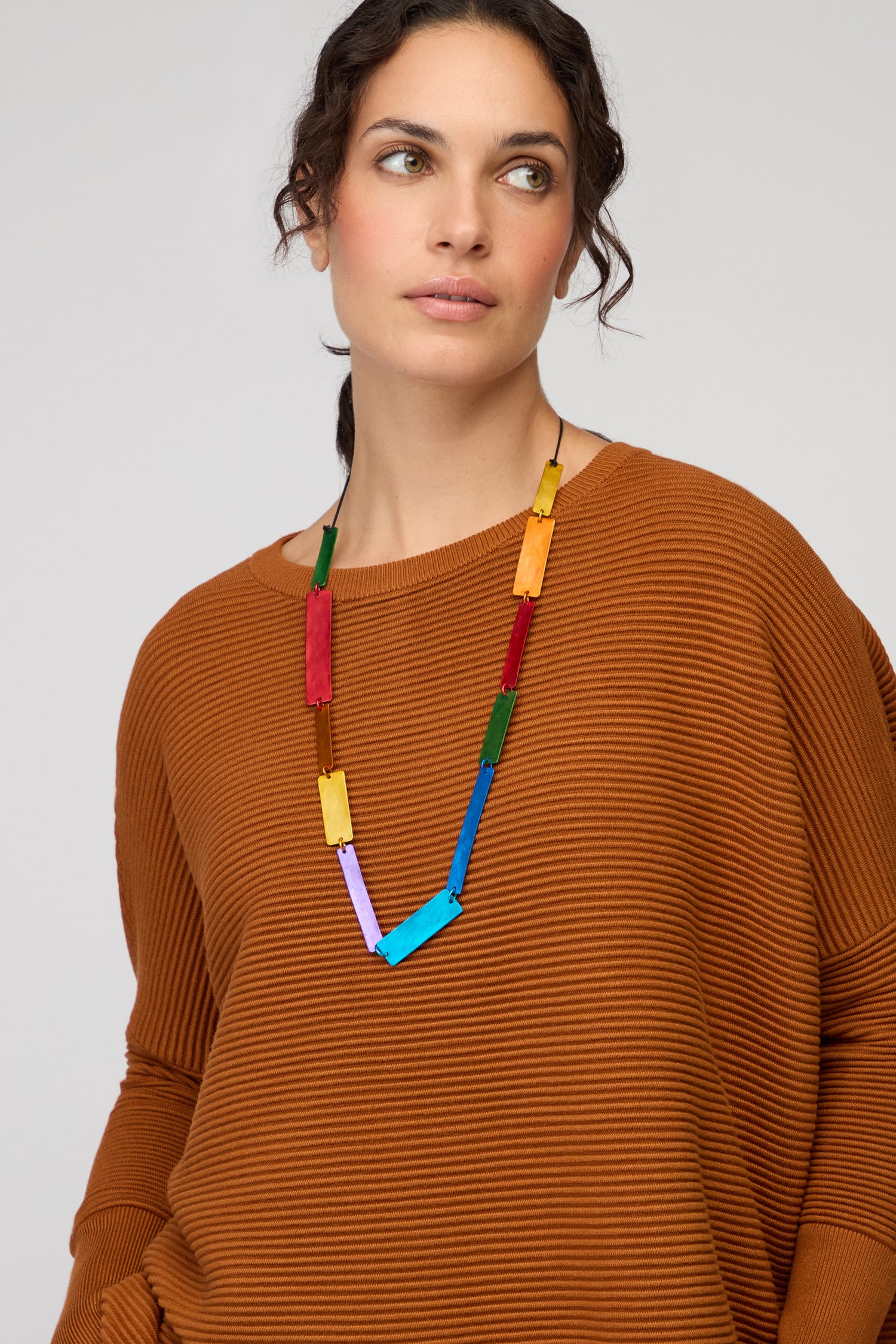 A woman in a rust-colored textured sweater wears the Colourful Rectangles Necklace—a long, colourful statement piece—while looking slightly to her left against a plain background.