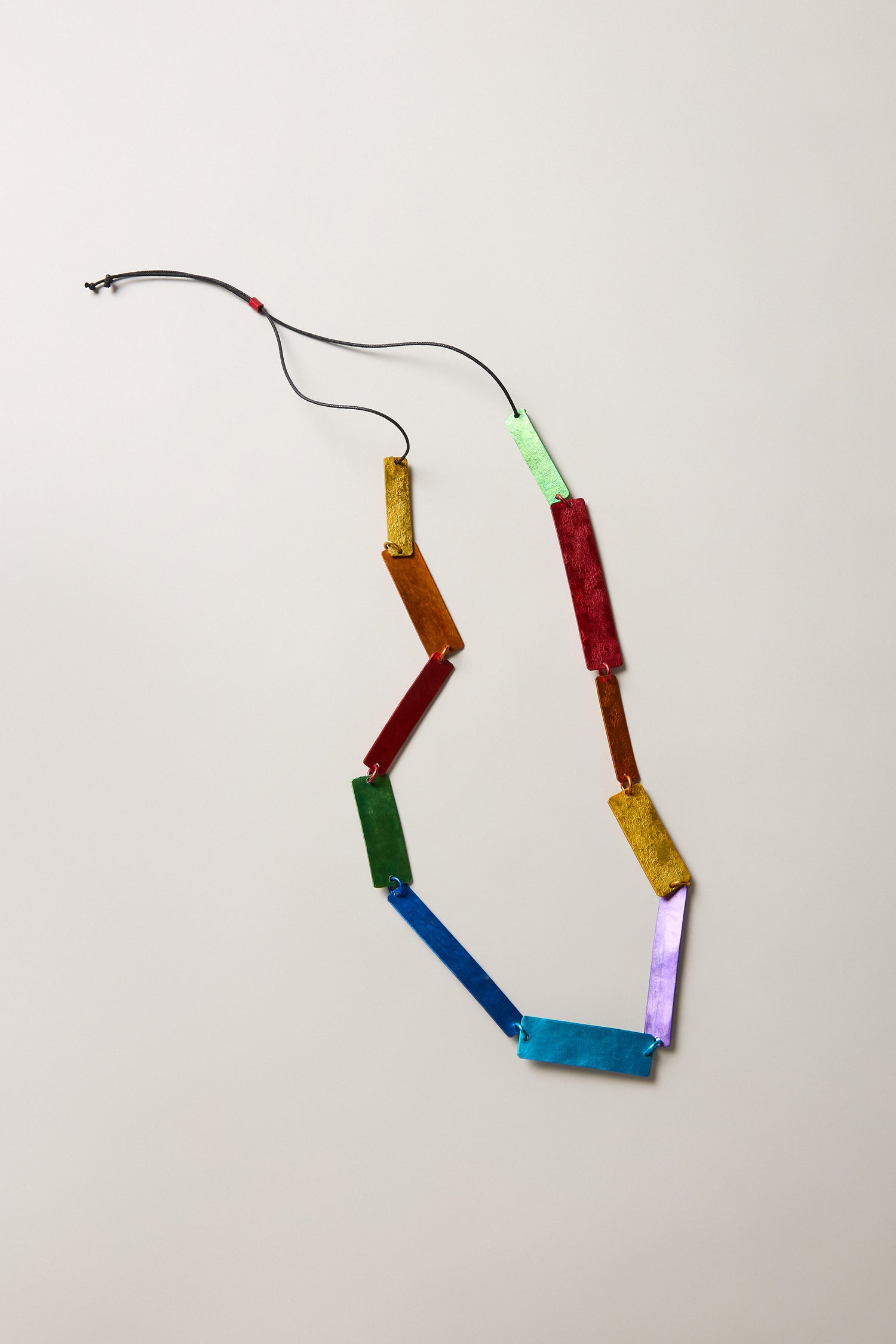 The Colourful Rectangles Necklace features multicolored rectangular links made from anodised aluminium, joined by a black cord and displayed on a light surface.