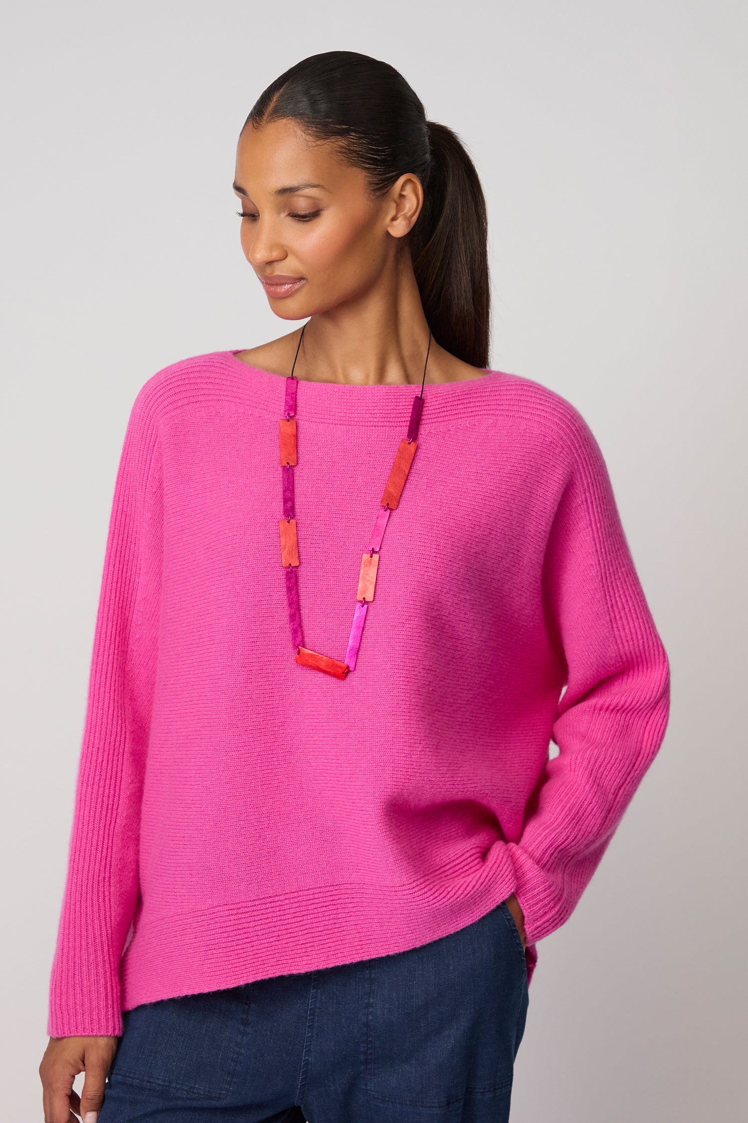 Woman wearing a bright pink sweater and the Colourful Rectangles Necklace, standing with one hand in her pocket and looking down.