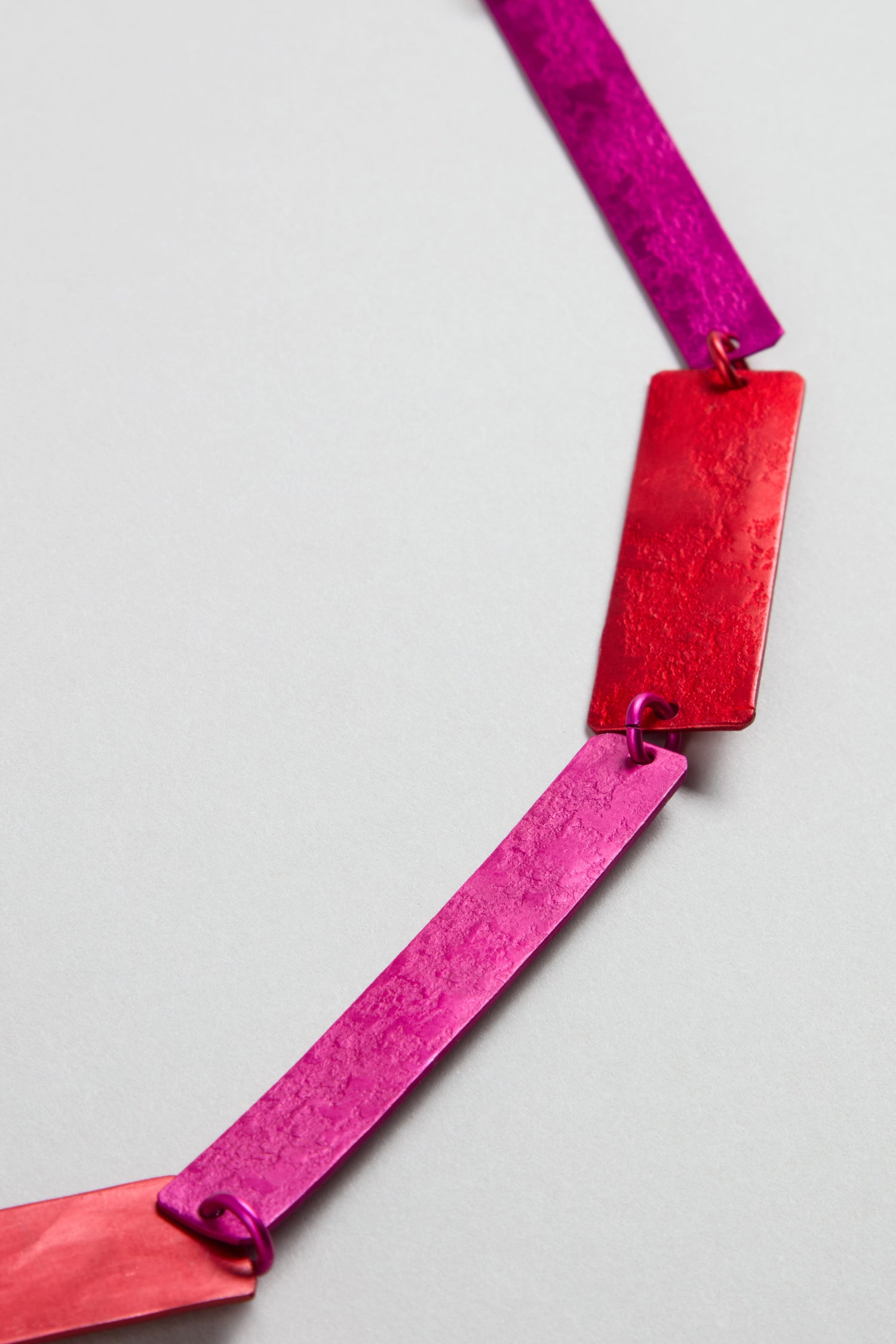 A close-up of the Colourful Rectangles Necklace, featuring rectangular anodised aluminium links in pink, magenta, and red joined by small rings, displayed on a light gray background.