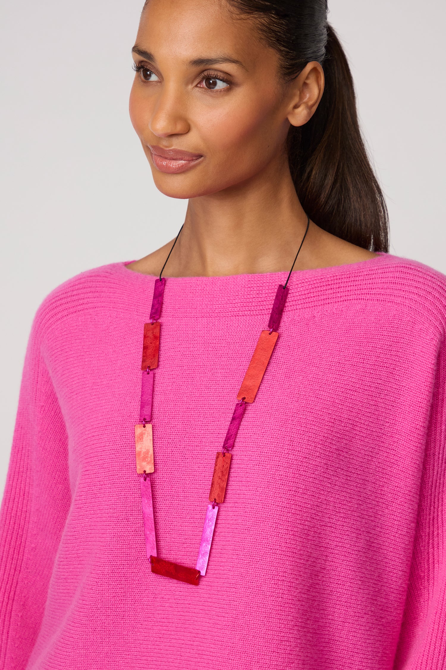 A woman wearing a bright pink sweater and the Colourful Rectangles Necklace, featuring long rectangular pink and red beads, looks slightly to the side.