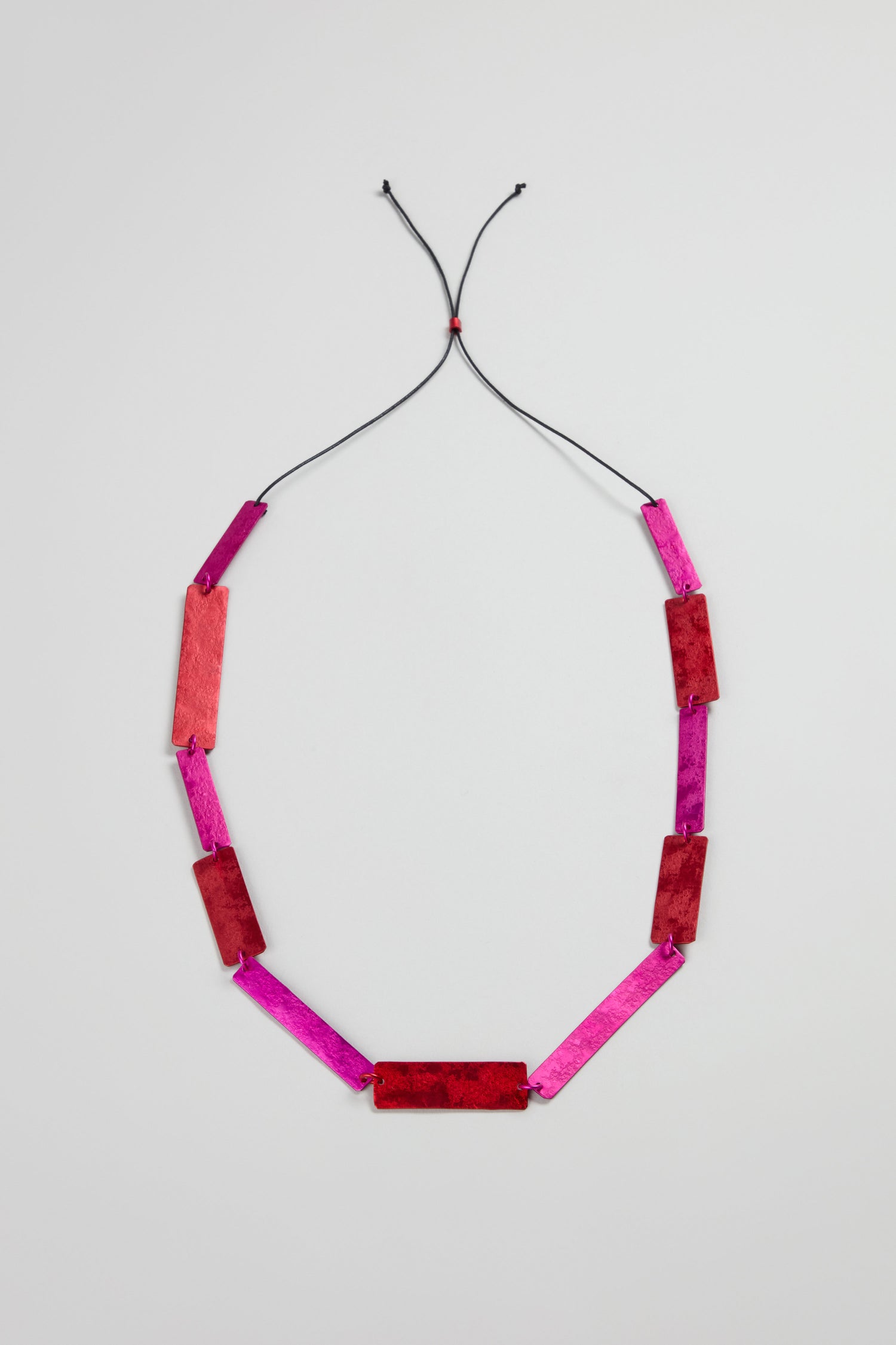 The Colourful Rectangles Necklace features red and pink rectangular anodised aluminium segments on a black adjustable cord, displayed flat on a light gray background.