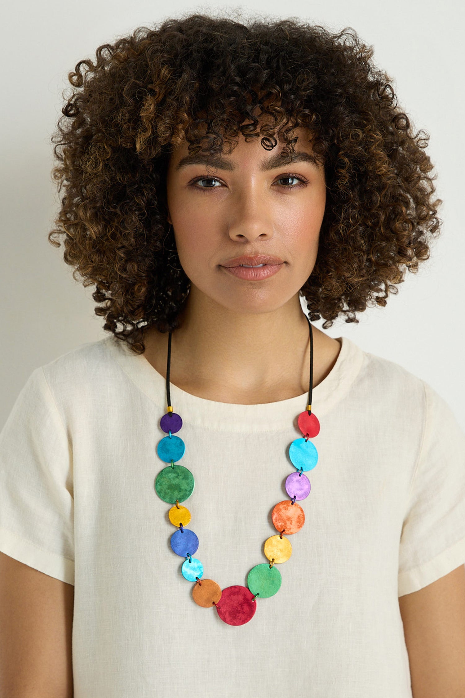 A woman with curly hair wears a cream top and the Colourful Aluminium Discs Necklace, featuring vibrant layered aluminium discs, against a plain light background.