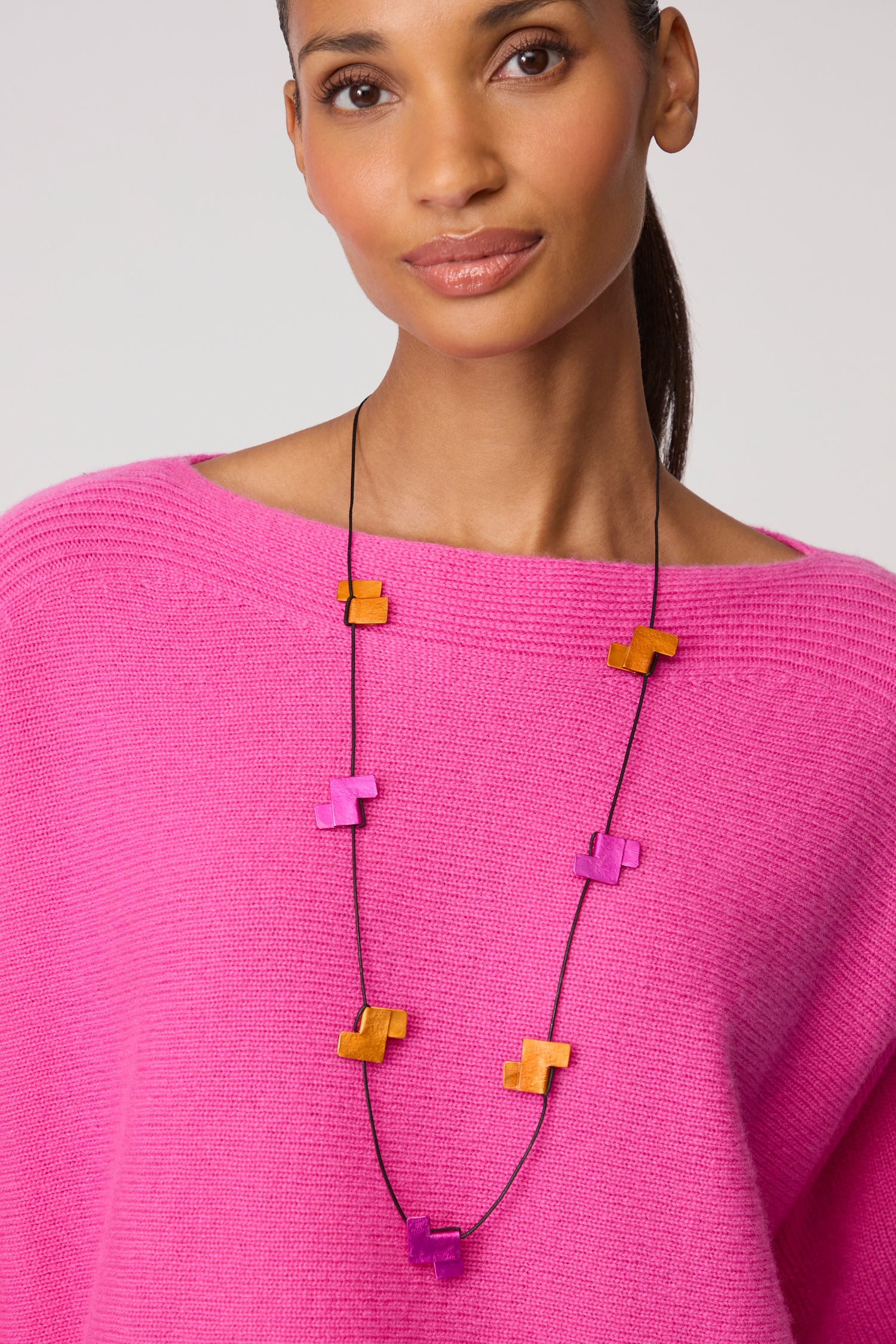 A woman wears a bright pink sweater and the Tetris Necklace, featuring alternating orange and purple geometric beads, for a bold contemporary jewellery look.