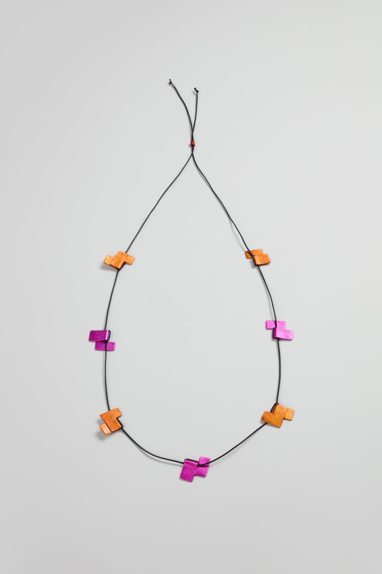 The Tetris Necklace is a black cord necklace with evenly spaced orange and magenta geometric wooden beads, showcasing a modern jewellery style against a simple light background.