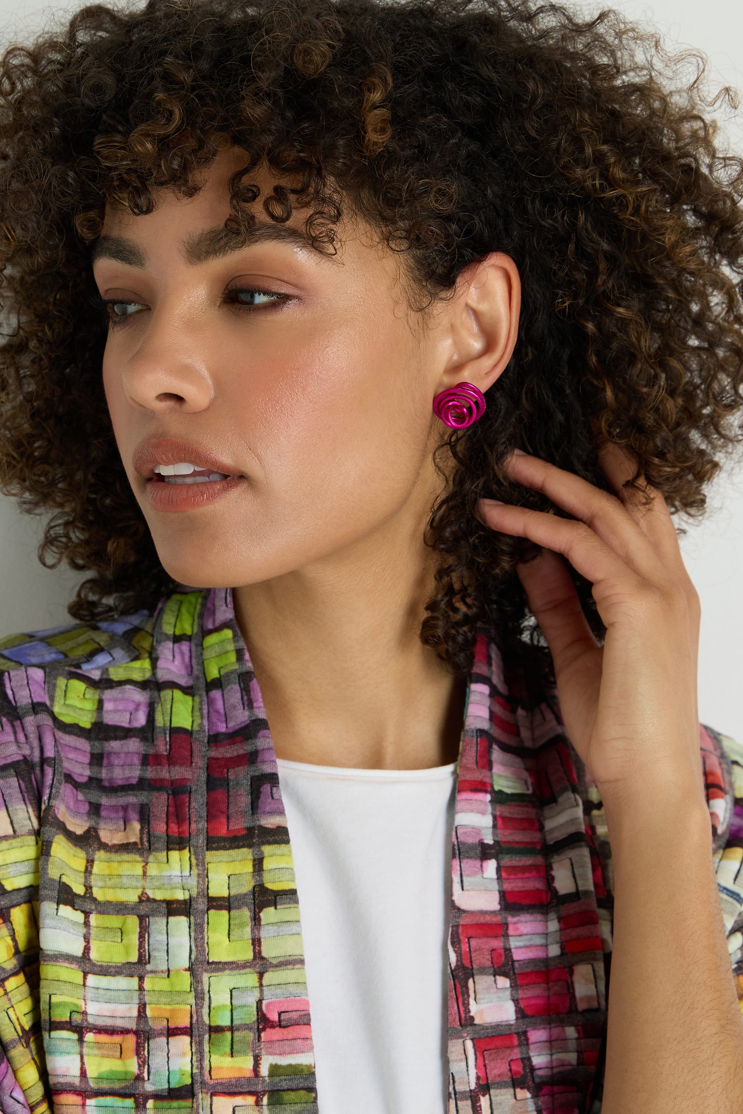 A woman with curly hair wears a colorful patterned jacket, a white top, and Mini Spiral Earrings in pink anodized aluminum shaped like roses, looking to the side while touching her hair.