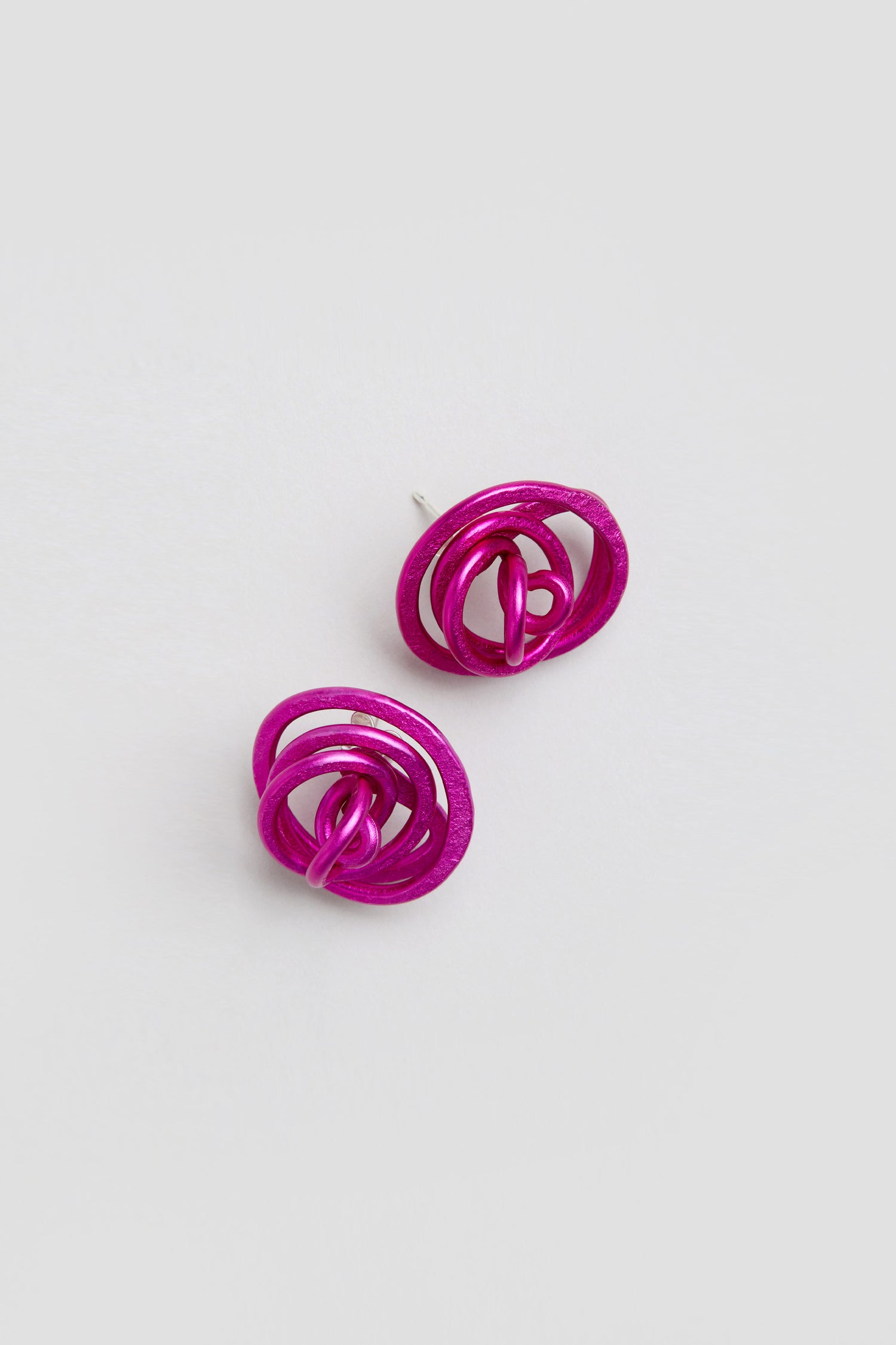 A pair of Mini Spiral Earrings, magenta metallic sculptural earrings made from anodized aluminium, displayed on a plain light gray background.