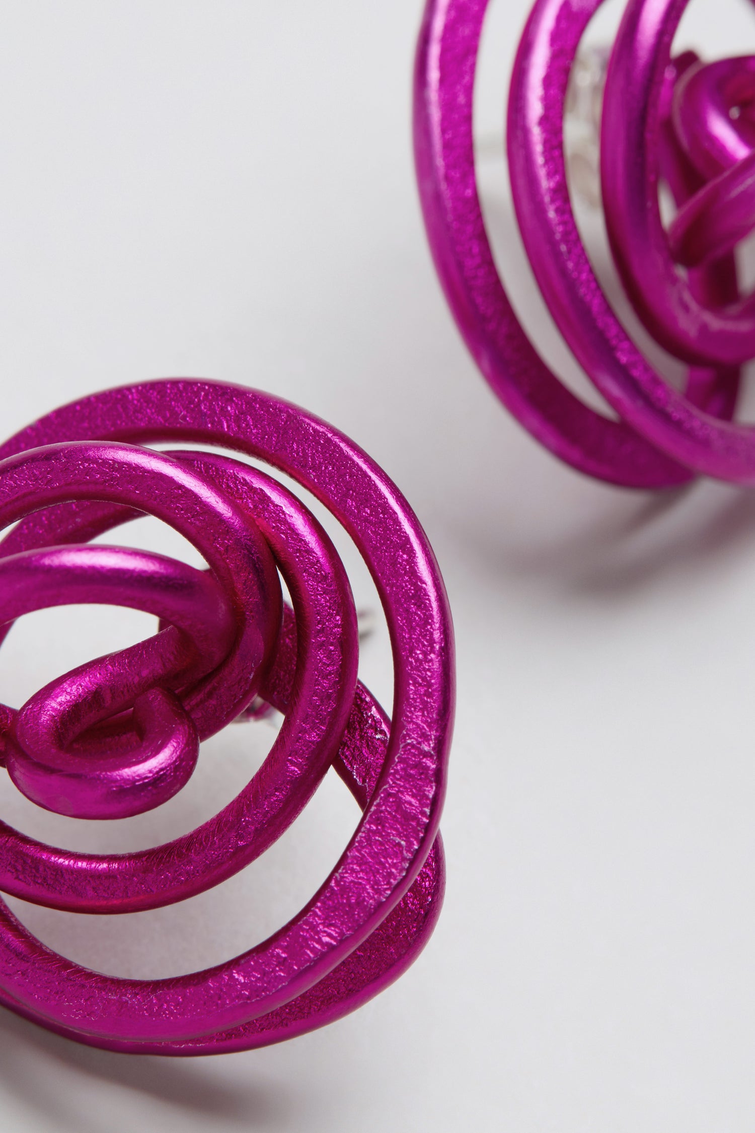 Close-up of two metallic pink Mini Spiral Earrings arranged on a white surface, highlighting their sculptural form and anodised aluminium finish.