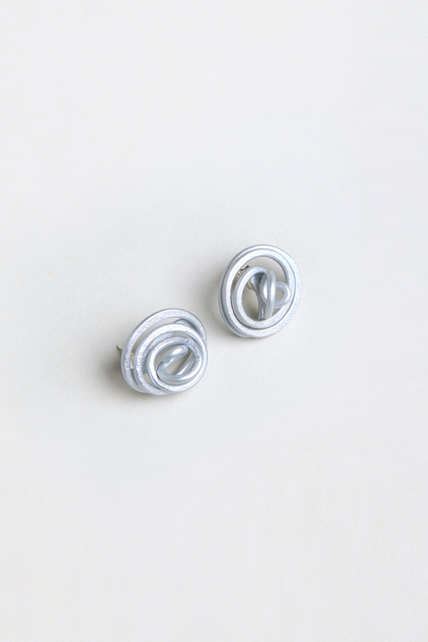 The Mini Spiral Earrings are sculptural silver earrings with interlocking circular loops, shown on a plain white background.