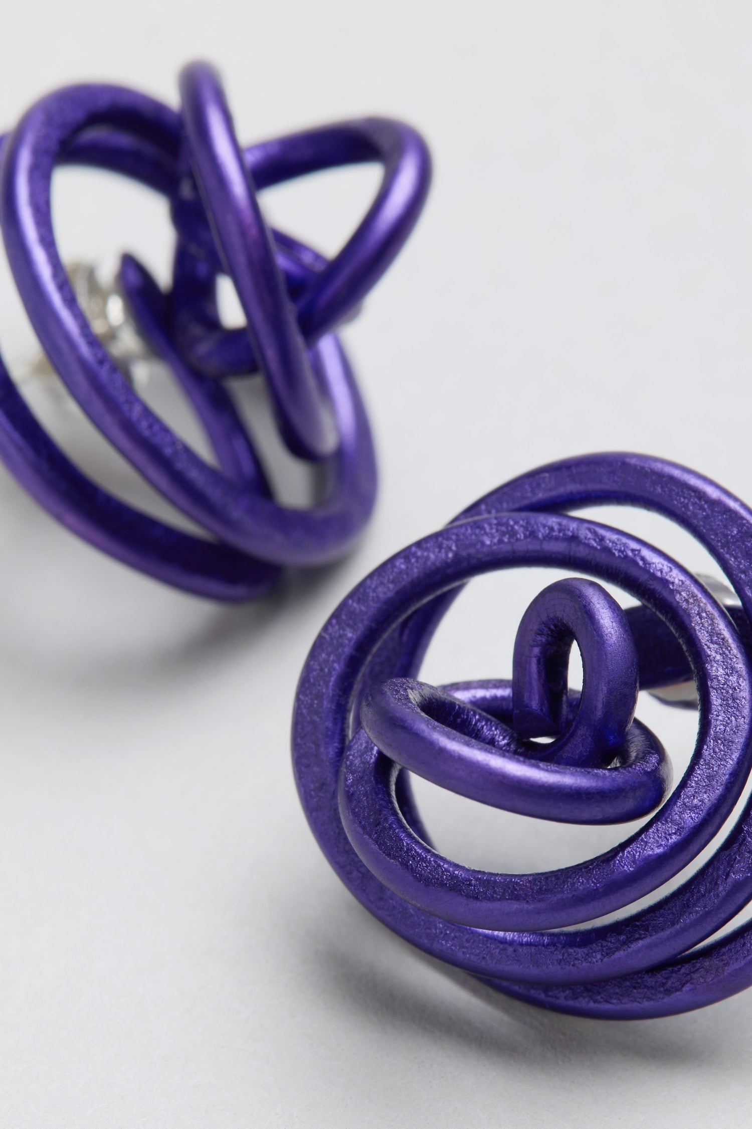 Close-up of two metallic purple Mini Spiral Earrings, sculptural and abstractly twisted, crafted from anodised aluminium and displayed on a light gray surface.