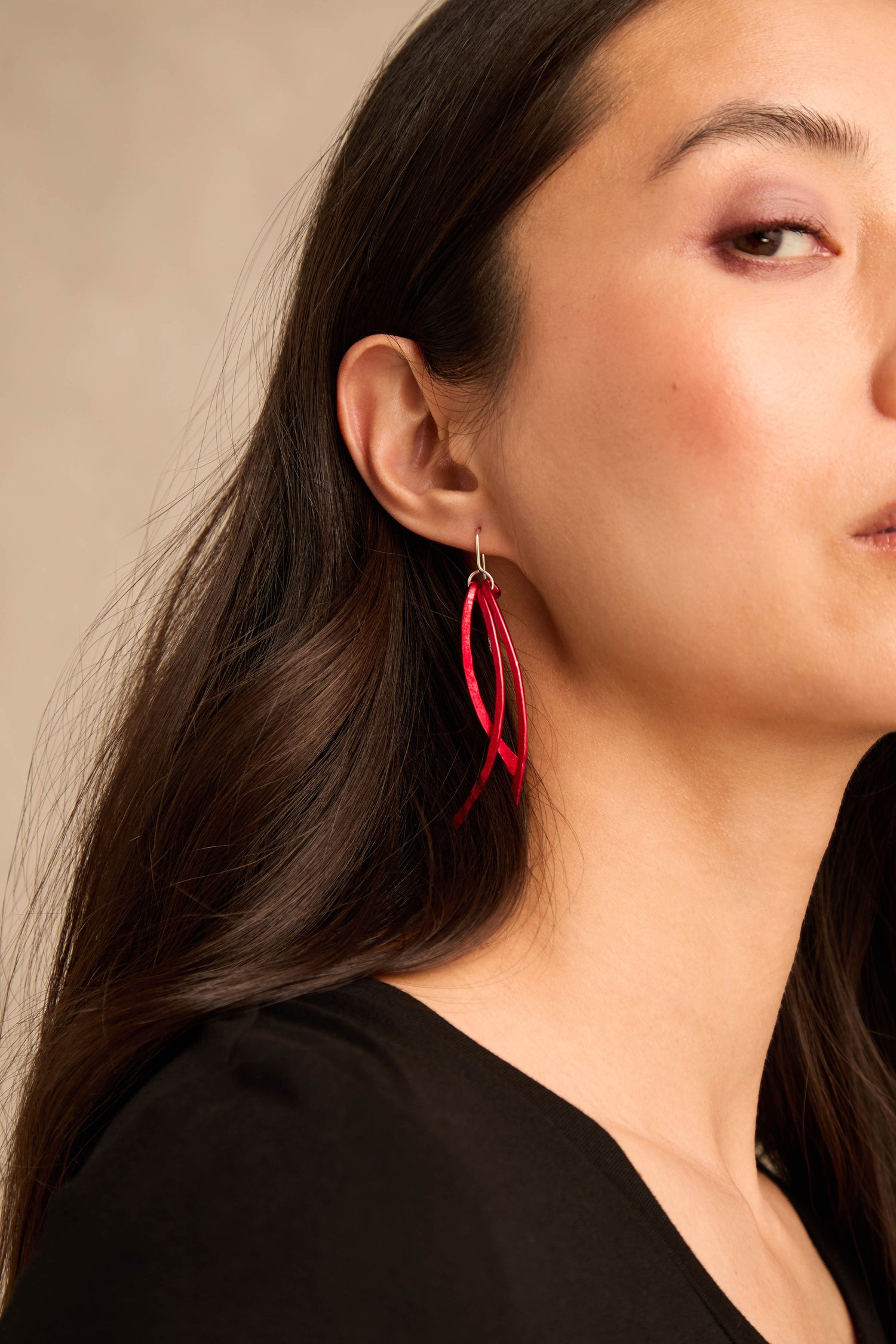 A woman with long dark hair, shown from the shoulders up in a neutral setting, wears a black top and the Arc Earrings—bold red anodised aluminium arcs with sterling silver hooks.