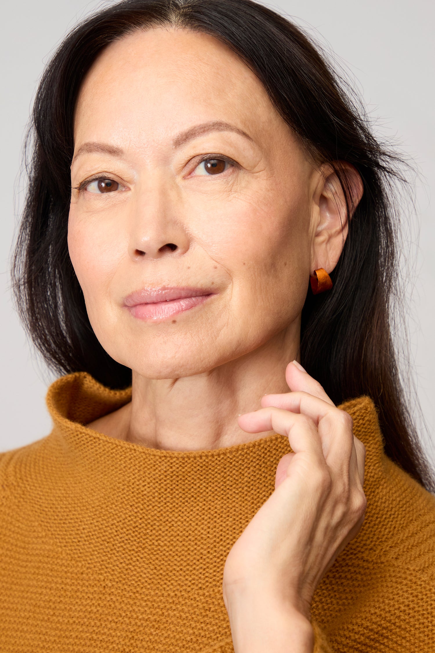 A woman with long dark hair wears a mustard knit top and Curved Earrings, gazing at the camera with a neutral expression and her hand near her face—an elegant showcase of contemporary jewelry.