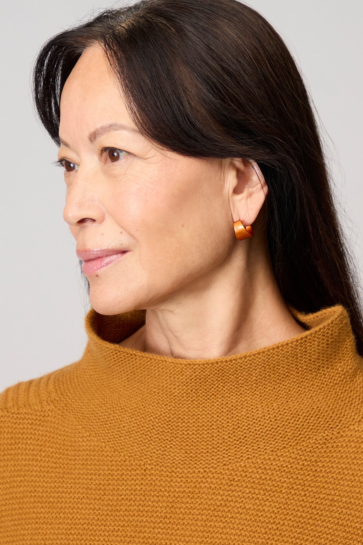 A woman with long dark hair, wearing a brown knit sweater and Curved Earrings, looks to the left against a neutral background—her statement accessories highlight her contemporary jewellery style.