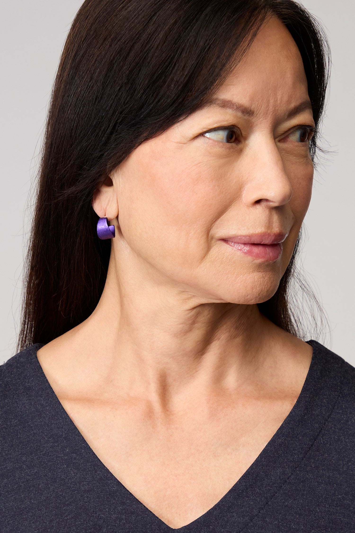 A woman with straight dark hair, wearing a navy V-neck top and Curved Earrings, looks to her right against a plain background—an elegant touch of contemporary jewellery.