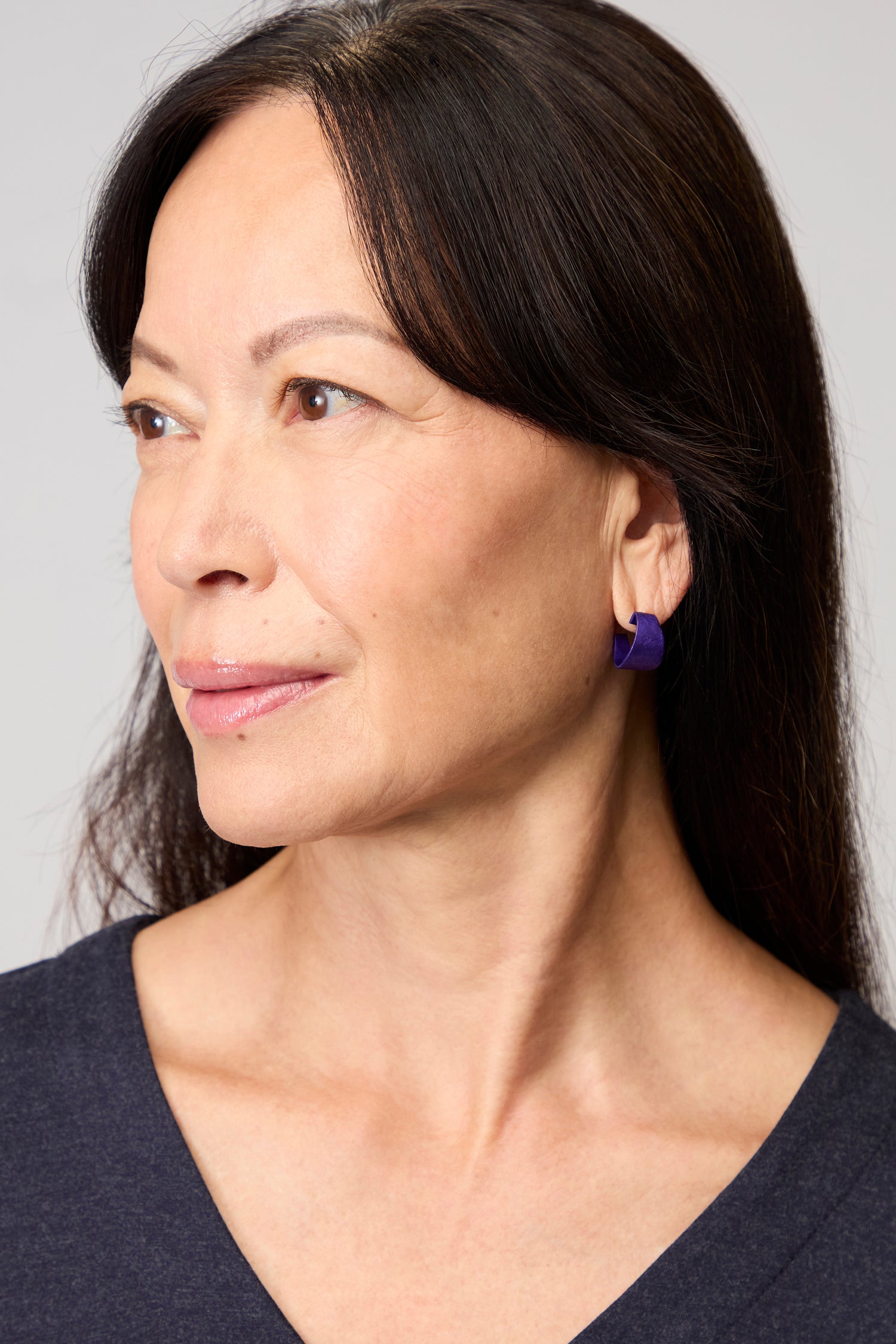 A woman with straight dark hair looks to the side, wearing a navy top and Curved Earrings—modern jewelry that adds a bold touch against a neutral background.