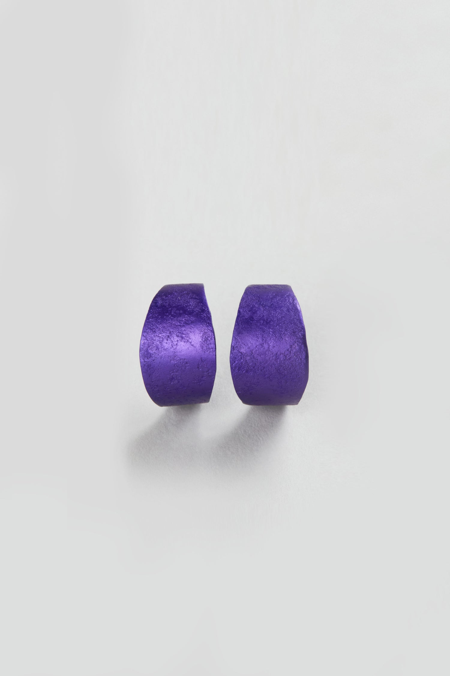 Two small, cylindrical pieces of purple material rest side by side on a plain white surface, resembling modern jewellery such as the Curved Earrings.