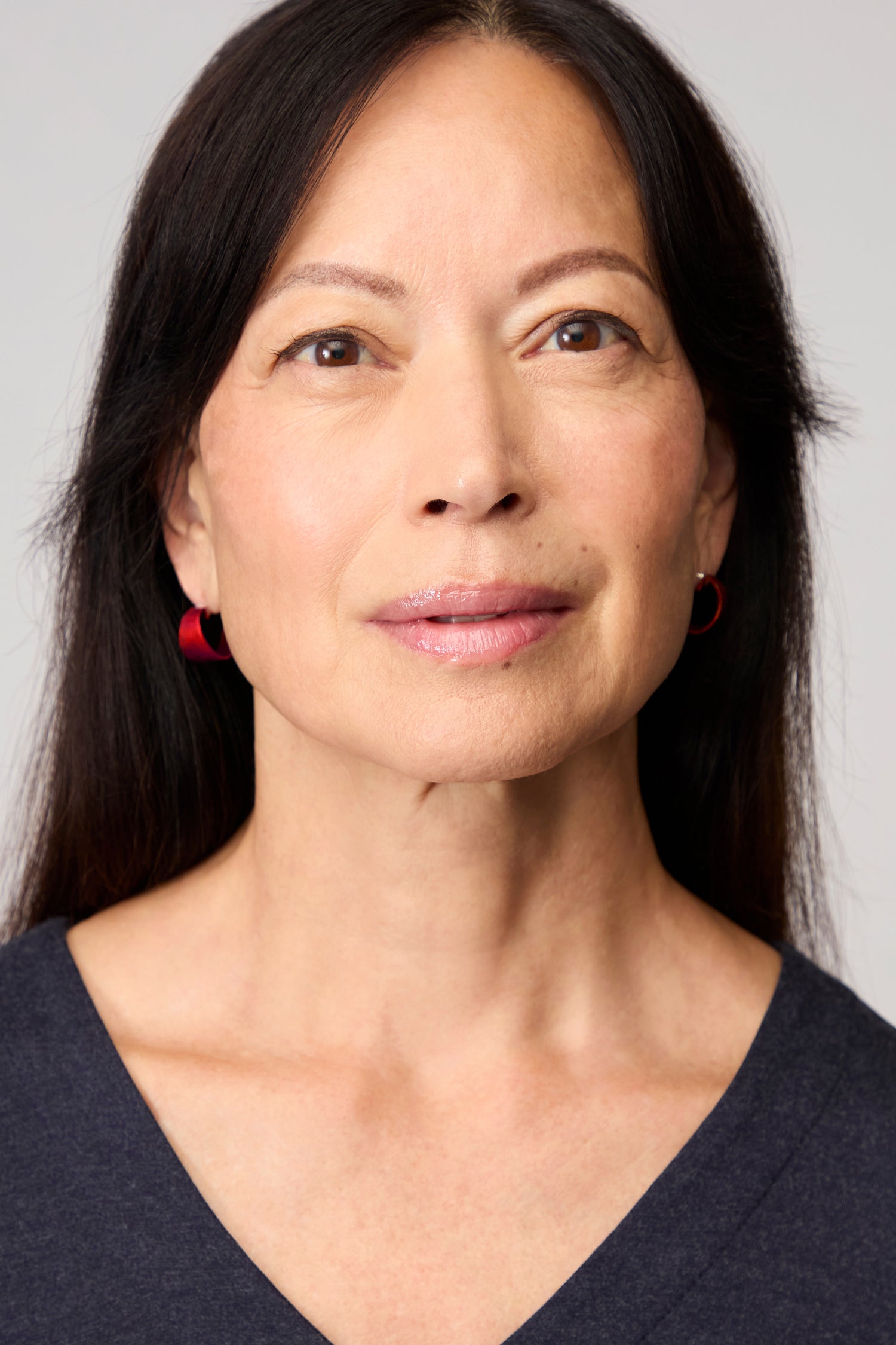 A woman with straight dark hair and red Curved Earrings gazes slightly upward, wearing a dark V-neck top that highlights the contemporary jewellery against a plain light background.