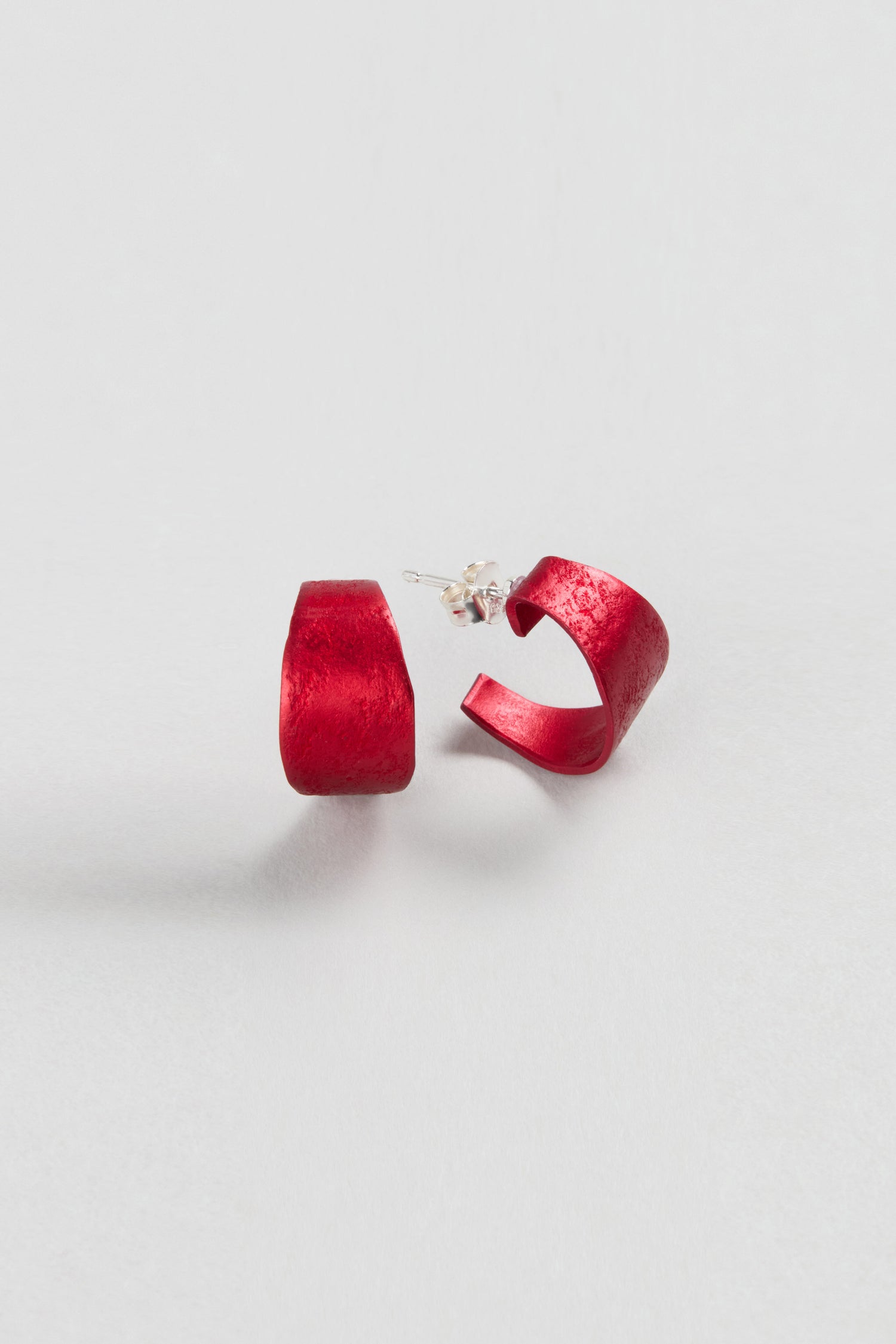 A pair of red Curved Earrings by Oana Millet with semi-circular design and silver posts, shown on a plain white background—ideal as contemporary jewellery and bold statement accessories.