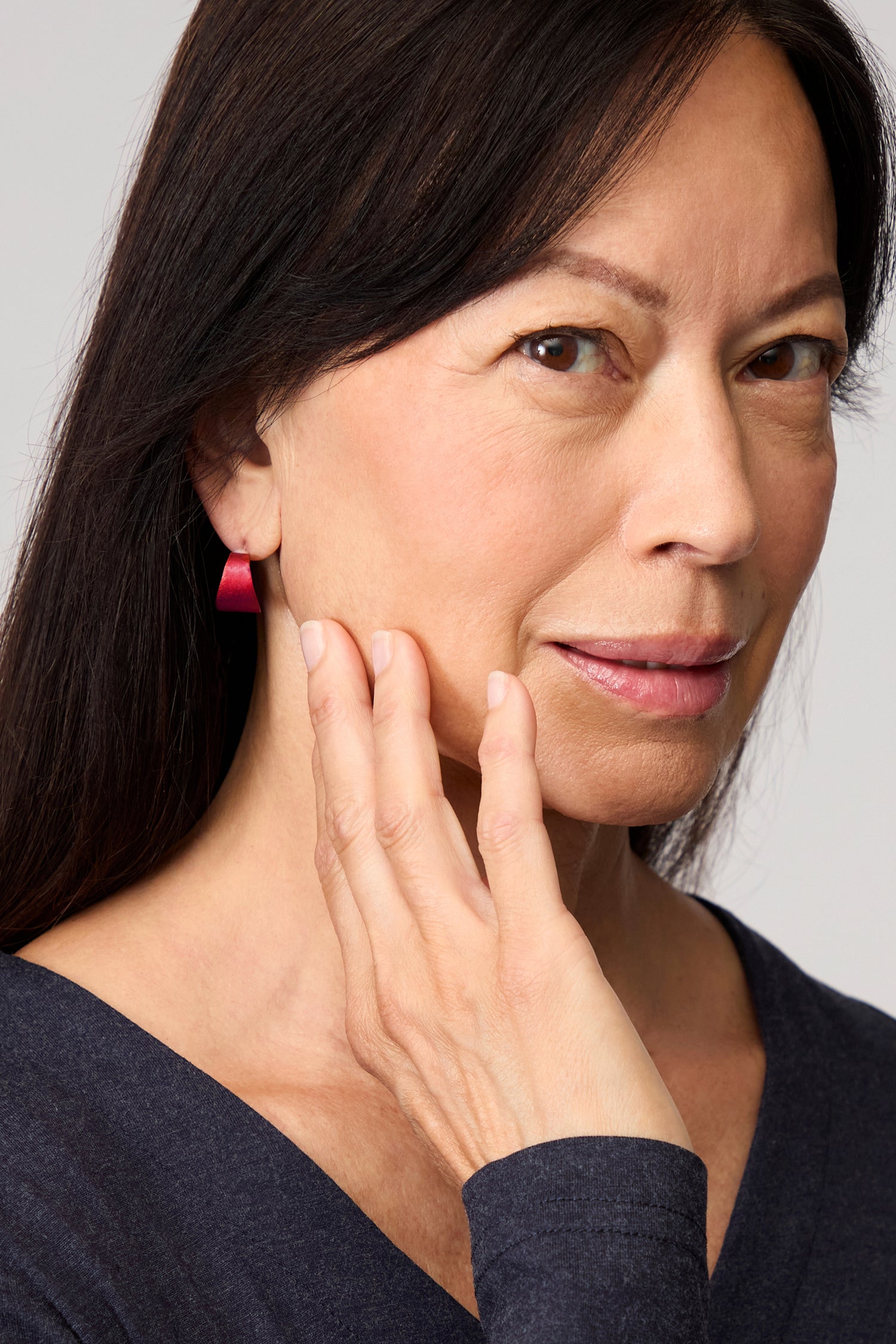 A woman with long dark hair wearing a dark top and bold pink Curved Earrings looks to her left against a neutral background.
