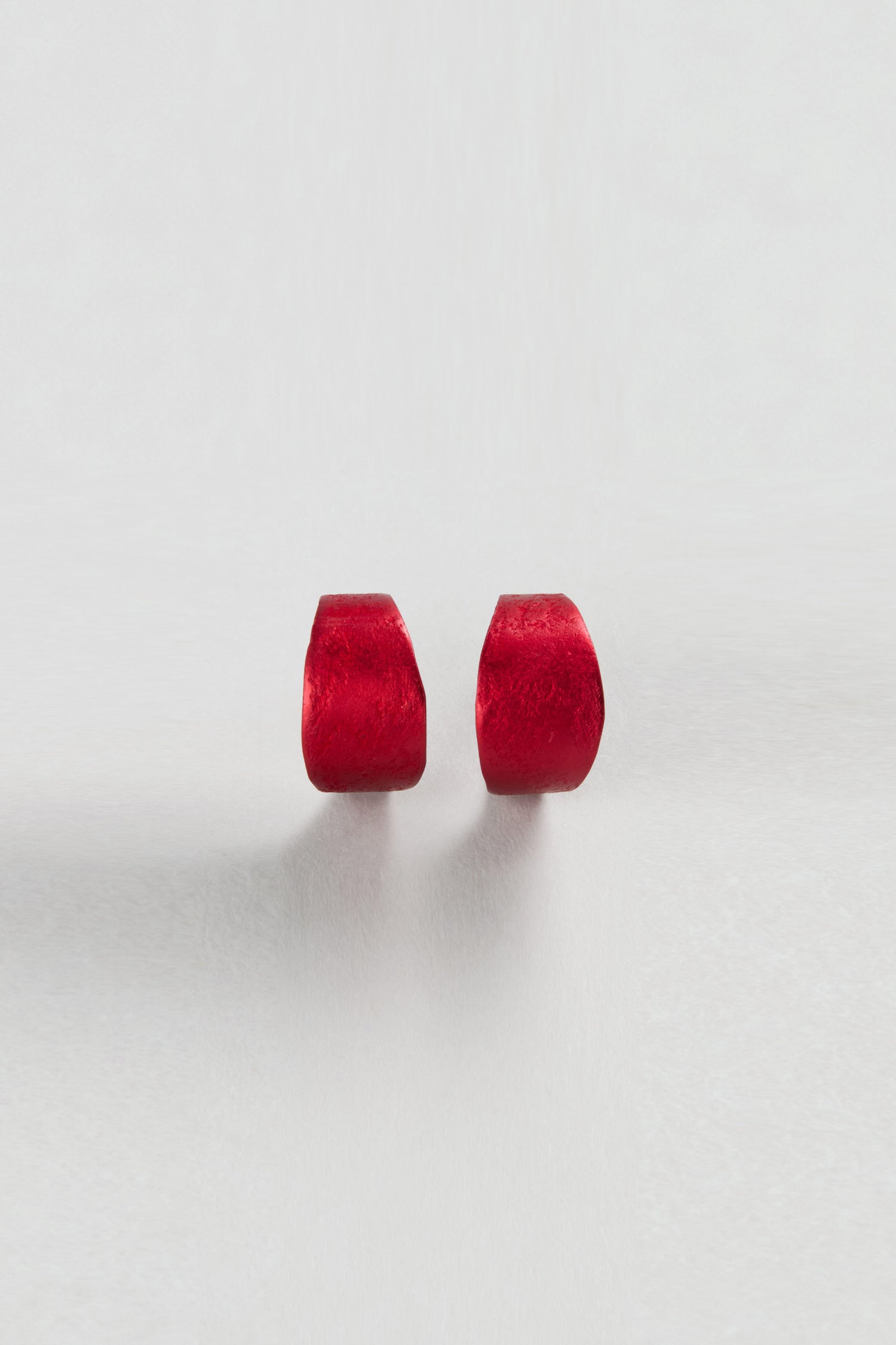 Two small, red, curved earrings are placed side by side on a light surface, echoing the modern style of Curved Earrings—ideal as unique statement accessories.