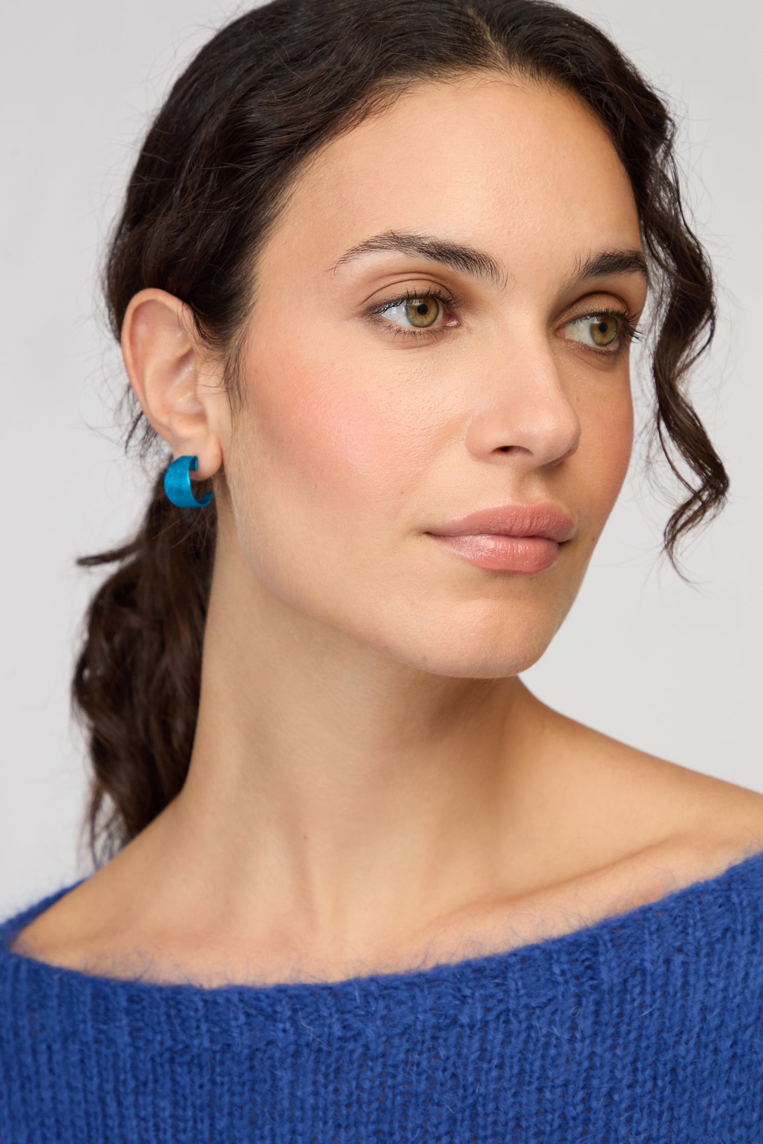Woman with wavy brown hair in a ponytail, wearing a blue sweater and the Curved Earrings, looking slightly to the side with a neutral expression—a modern look highlighted by contemporary jewelry.