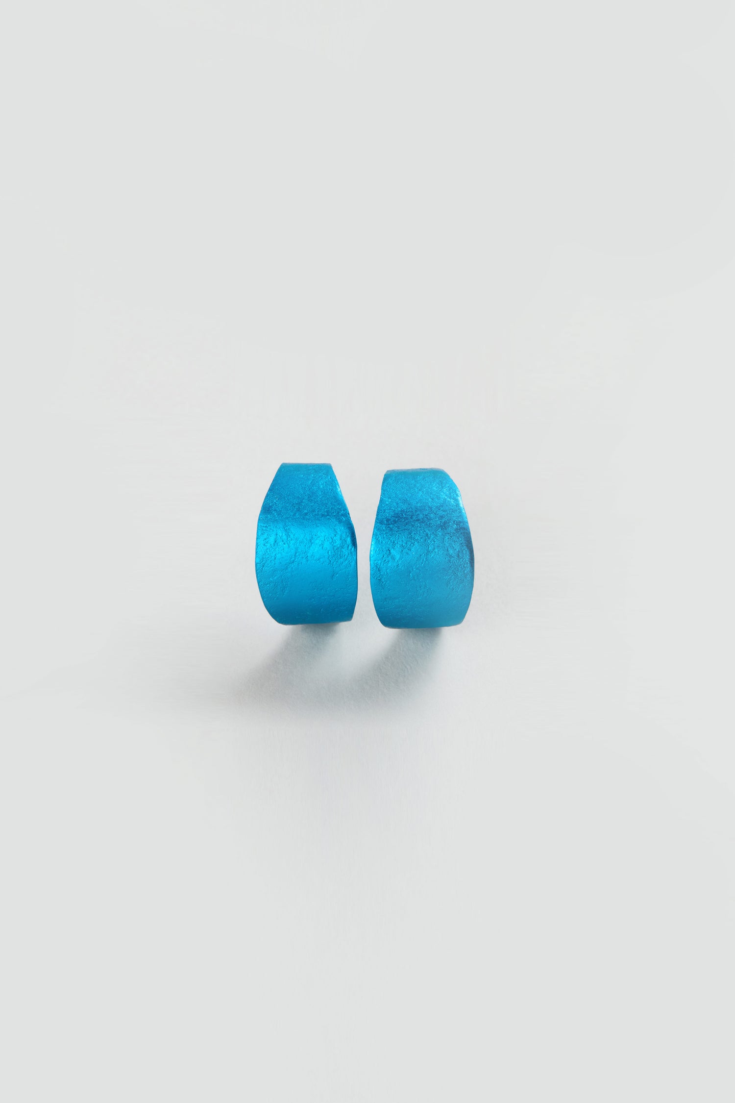 Two small, blue Curved Earrings with flat ends rest side by side on a plain white background, capturing the modern elegance of minimalist statement jewellery.