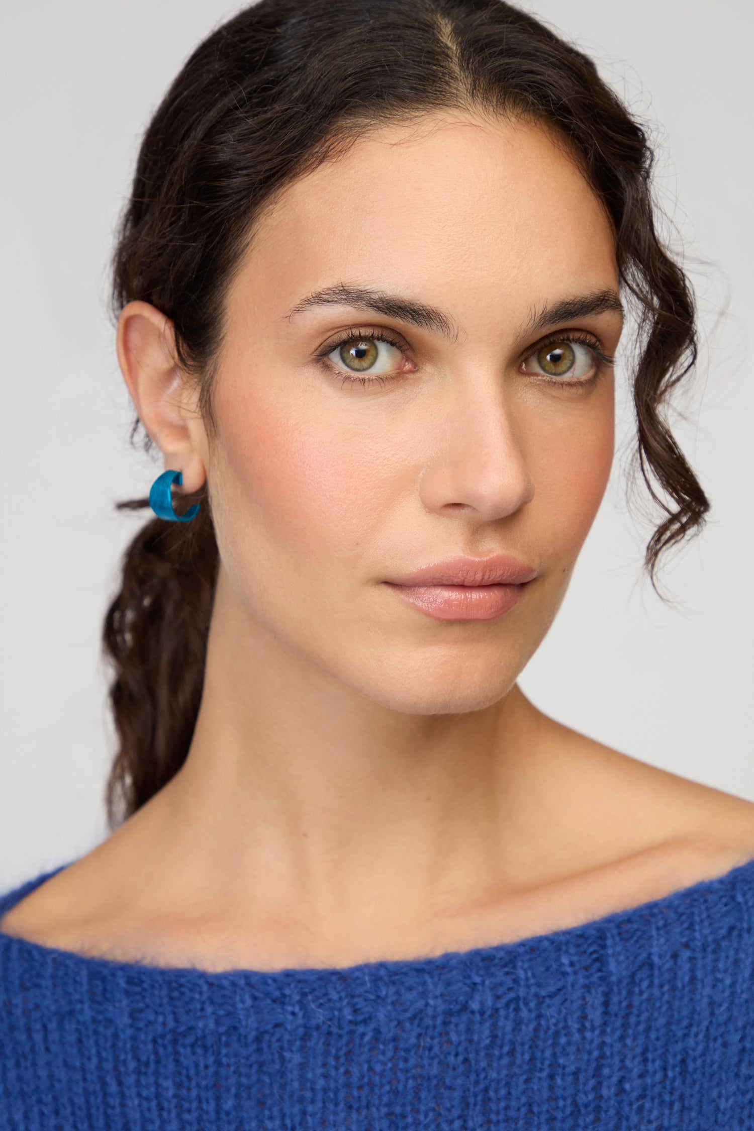 A woman with wavy dark hair, wearing Curved Earrings and a blue knit sweater, looks directly at the camera against a plain background.