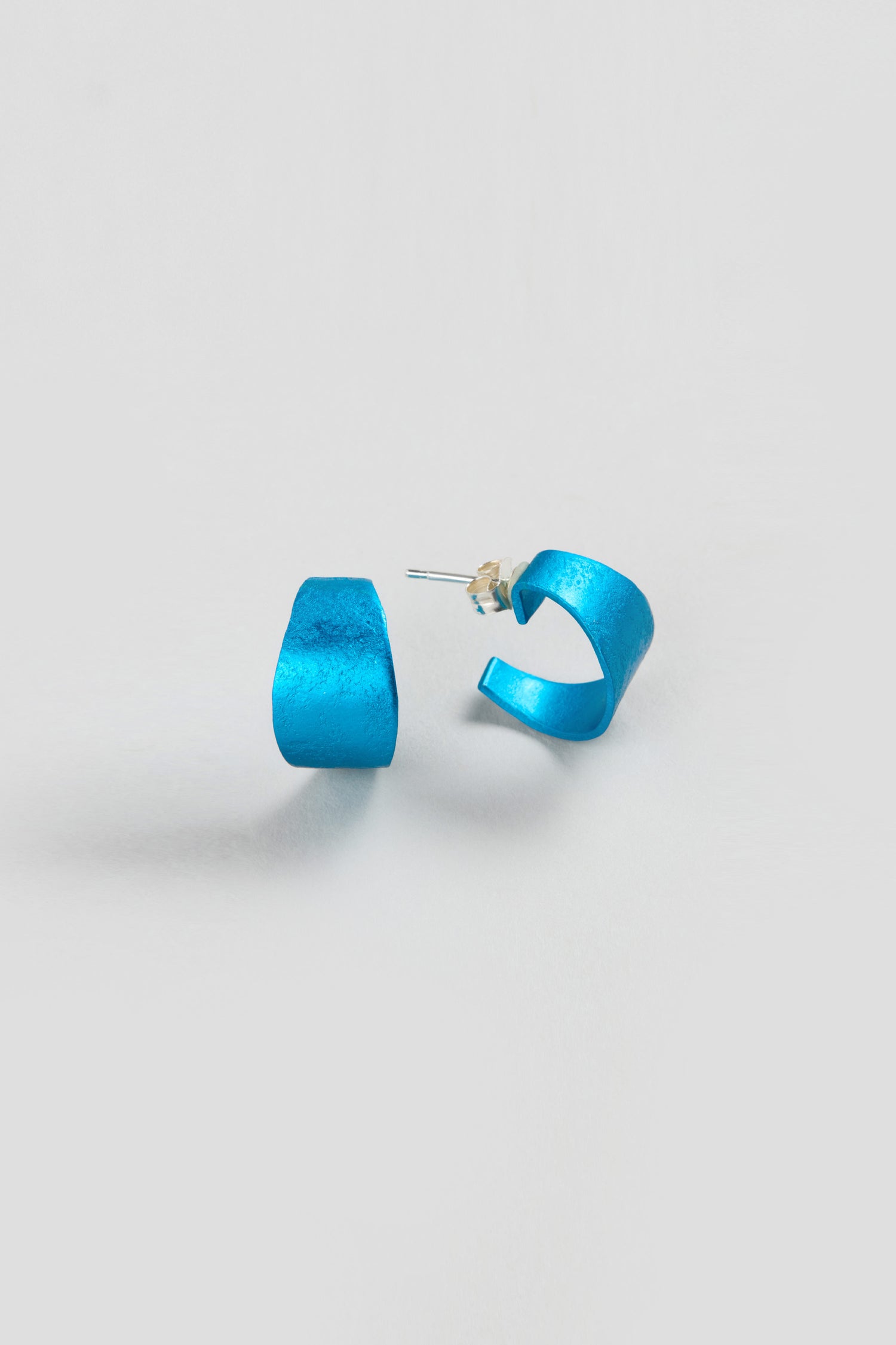 A pair of small, textured blue Curved Earrings with silver posts and clear backings, ideal for fans of contemporary jewellery, shown on a plain white background.