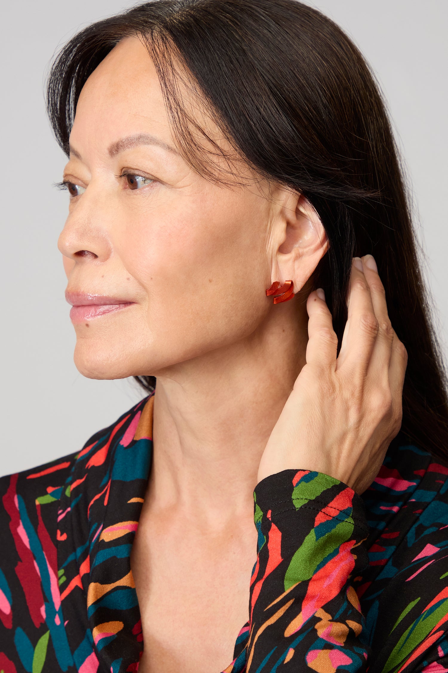 A woman with long dark hair looks to the side, touching her ear and wearing a colorful patterned top and striking Ribbon Earrings crafted from anodised aluminium.
