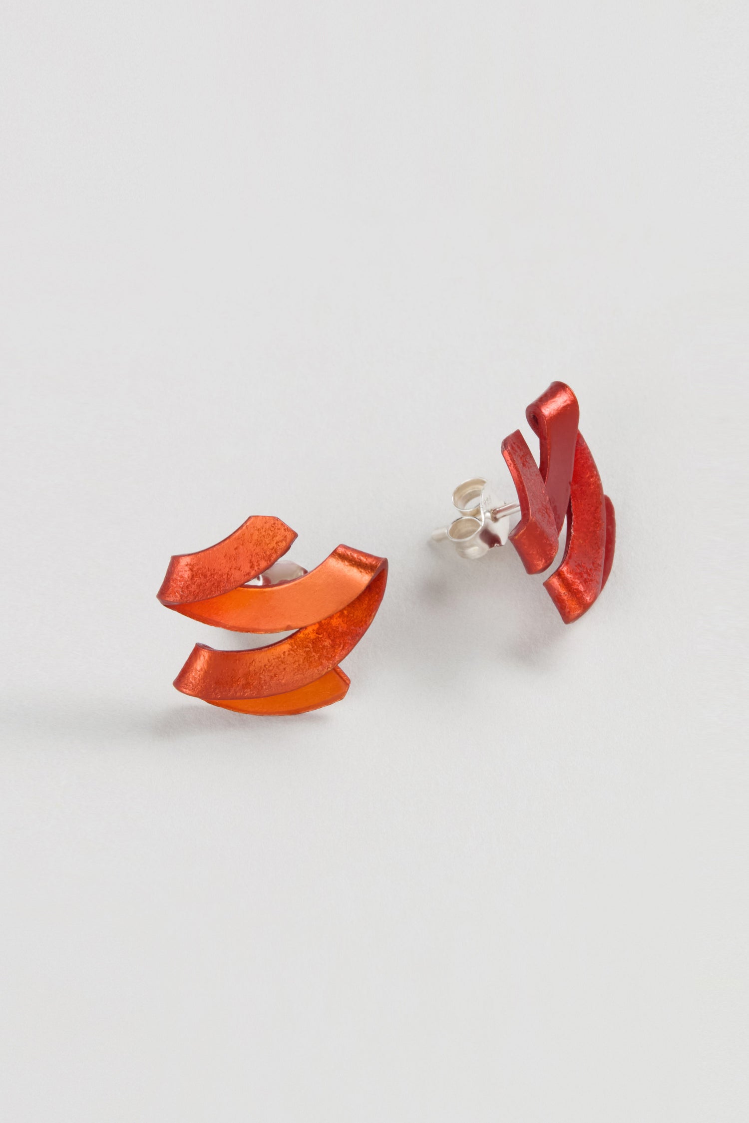 The Ribbon Earrings, made of red anodised aluminium in an abstract shape, are displayed on a plain white background, highlighting their sculptural jewellery design.
